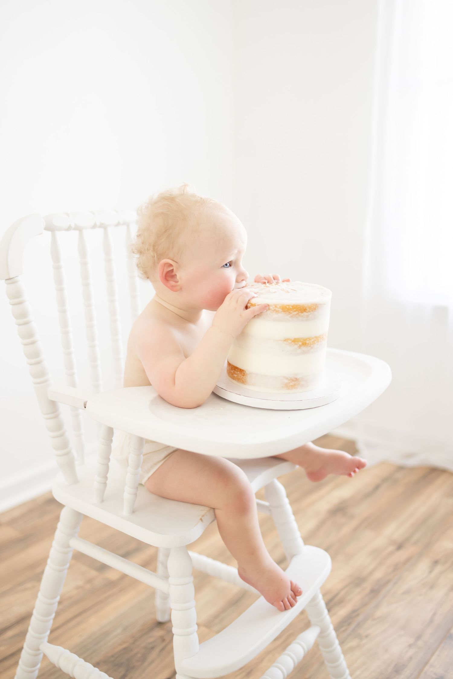 Adorable baby in diaper sitting in white high chair eating first birthday cake in bright white room.