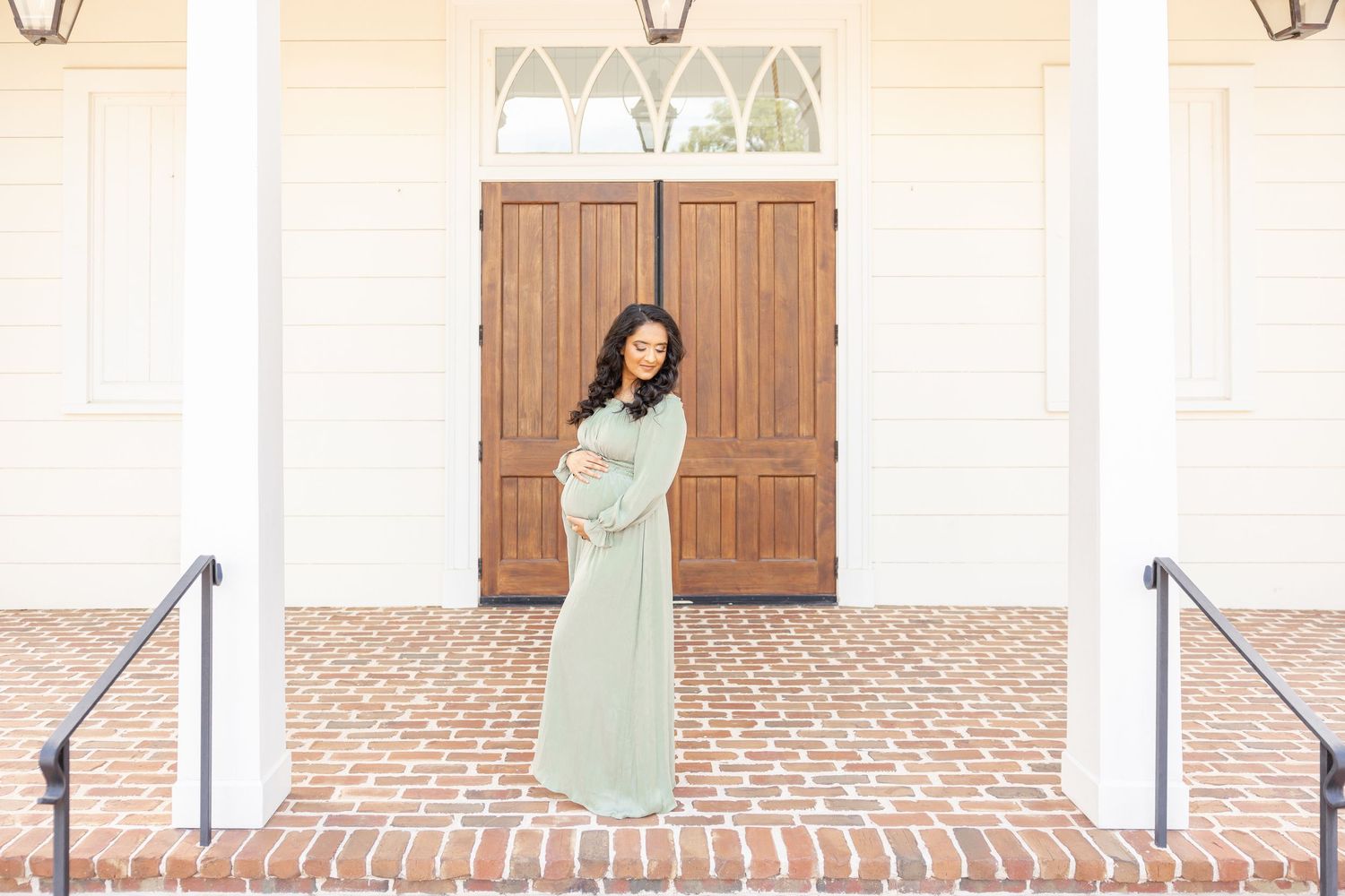 Pregnant woman in sage green maternity gown standing on brick steps before elegant white building with wooden doors.