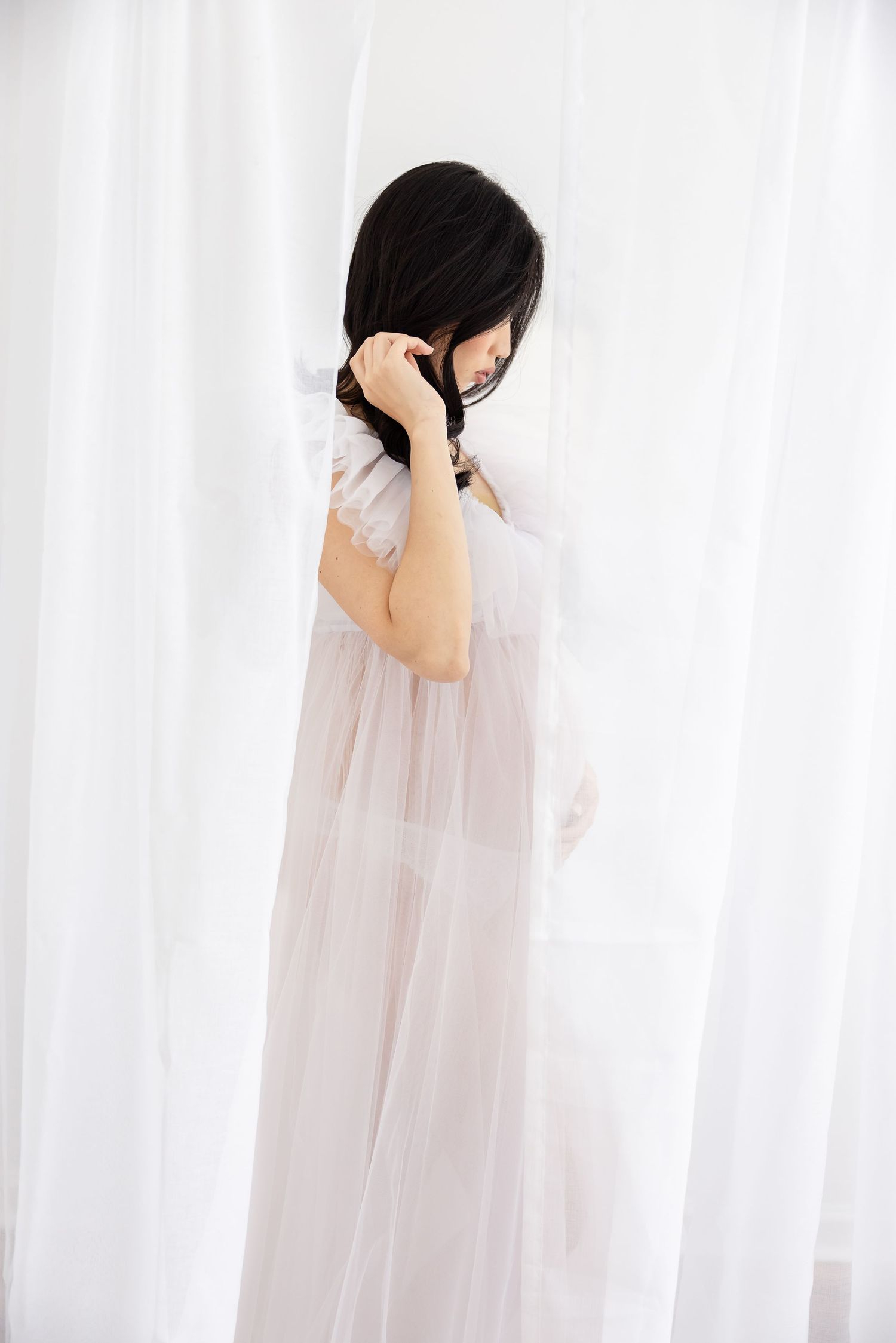 A woman in a sheer white dress stands gracefully against flowing white curtains, her dark hair cascading forward.