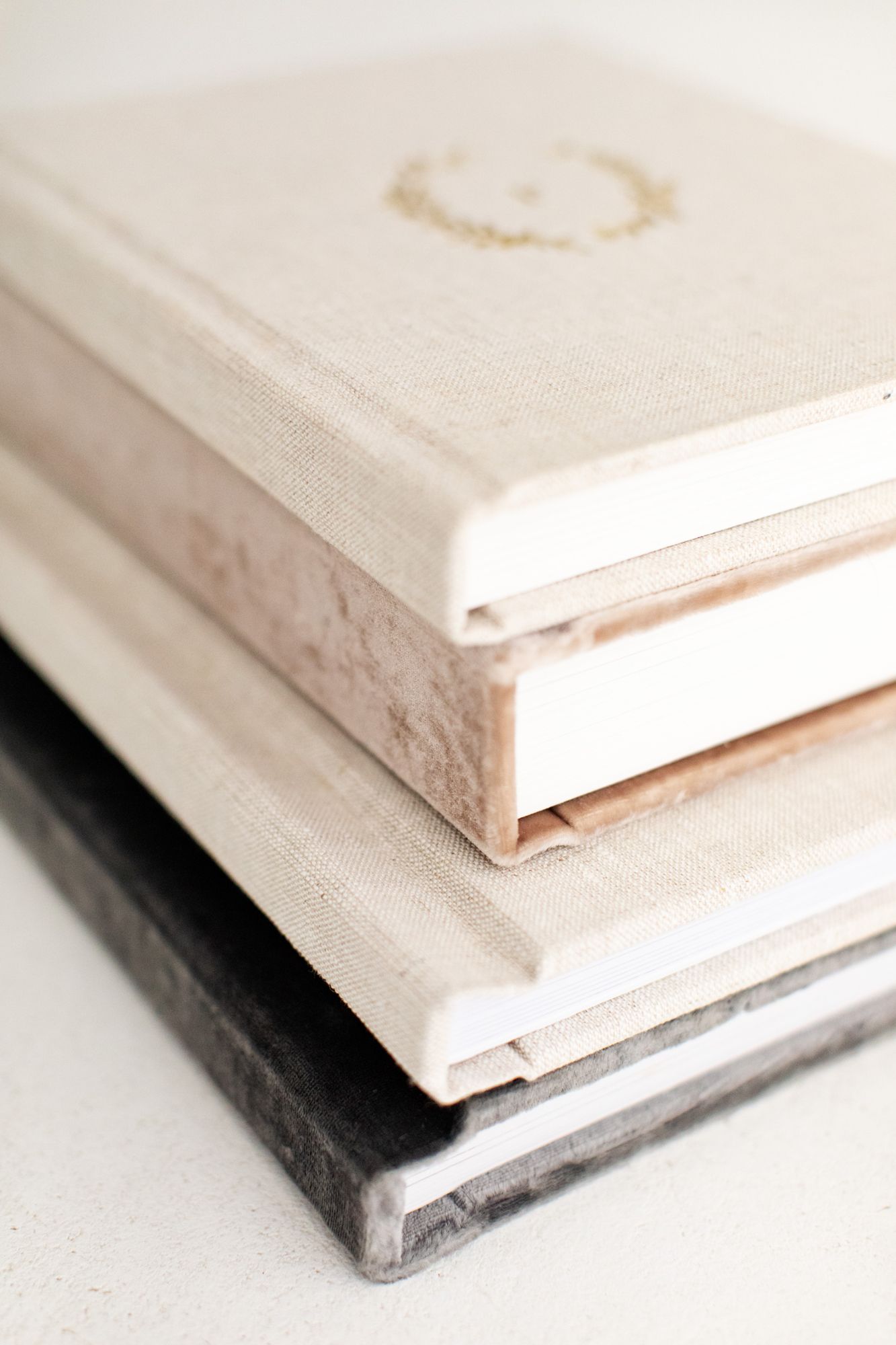 Stack of three elegant hardcover books in neutral tones, featuring linen and velvet textures on a white surface.