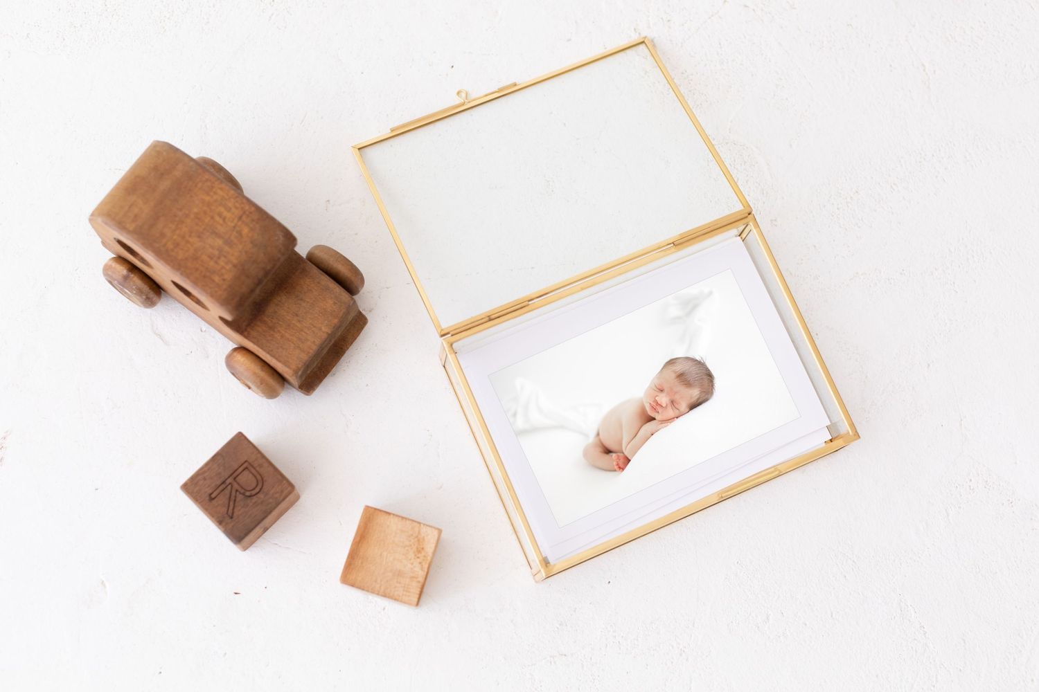 Gold glass keepsake box with newborn photo print, wooden toy car and blocks on white background.