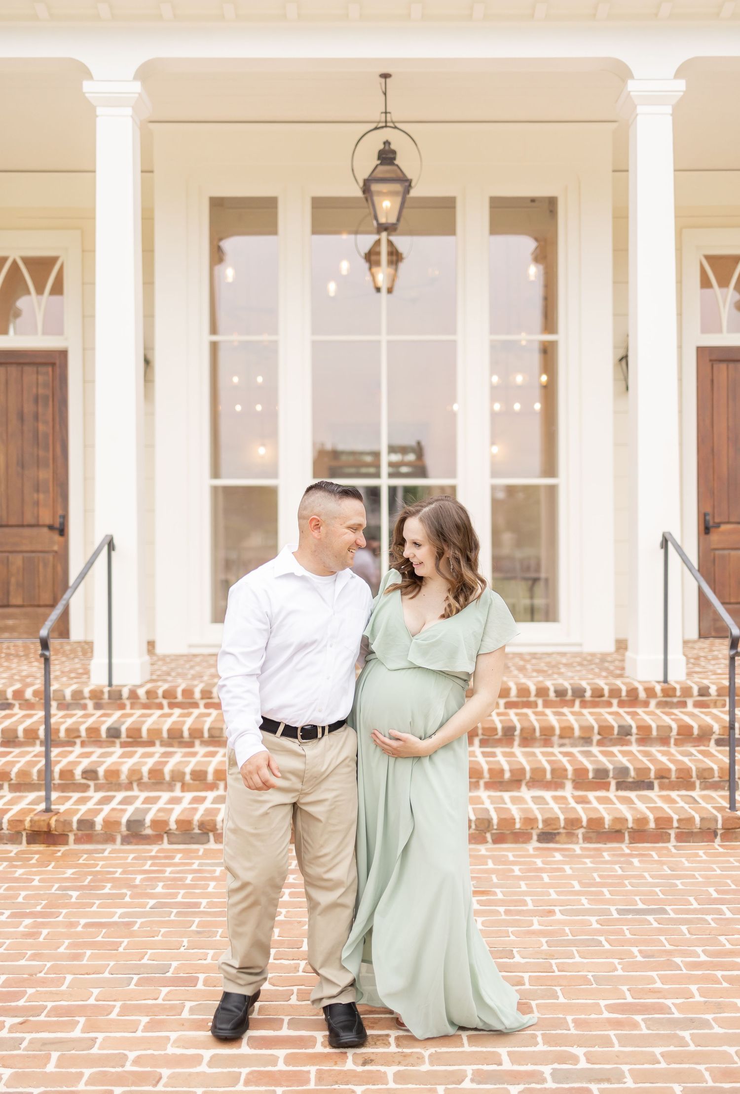 Couple posing for maternity photos on brick steps of elegant white building, woman in sage green gown cradling baby bump.