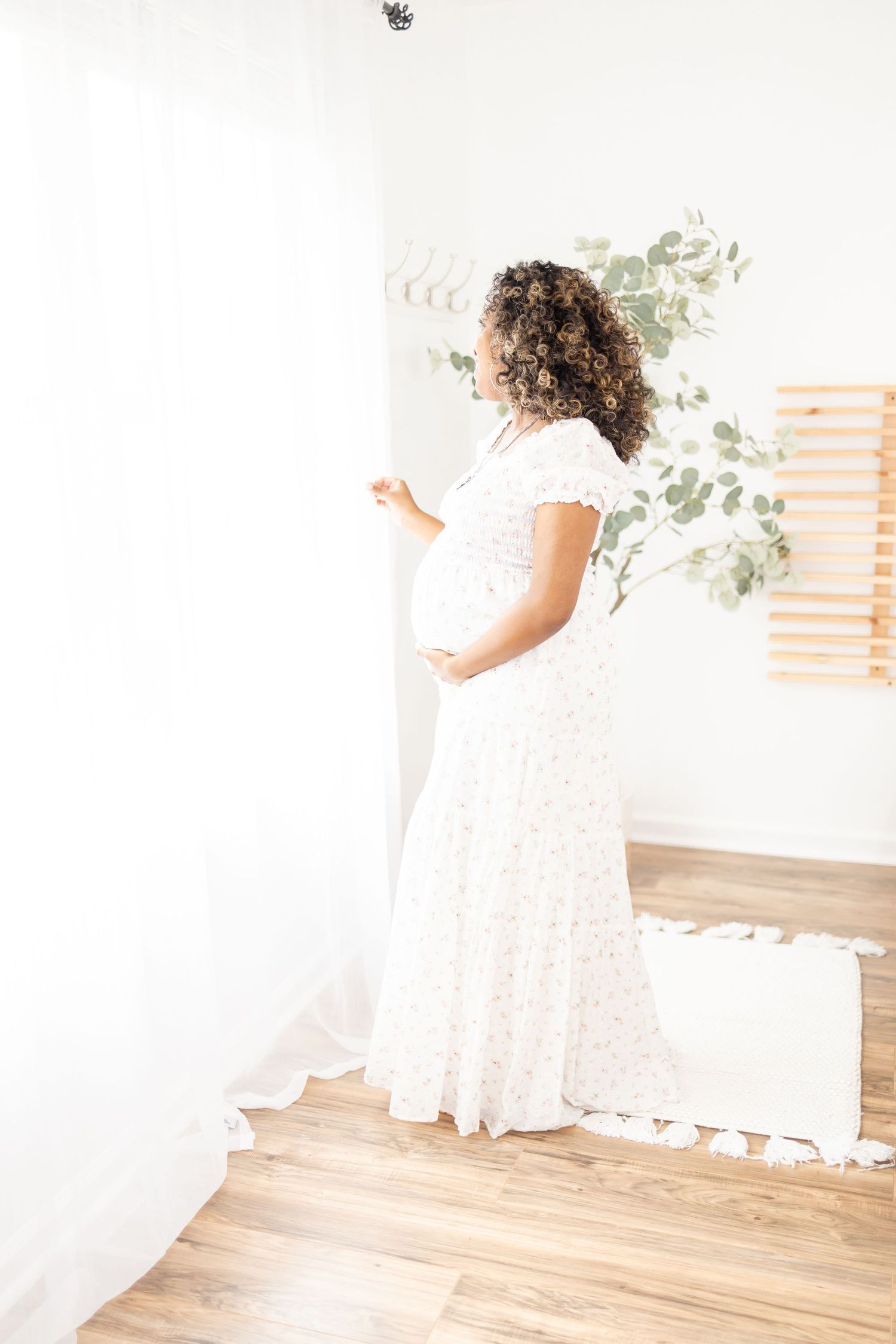 A pregnant woman in a flowing white lace dress stands beside white curtains and decorative eucalyptus in a bright room.