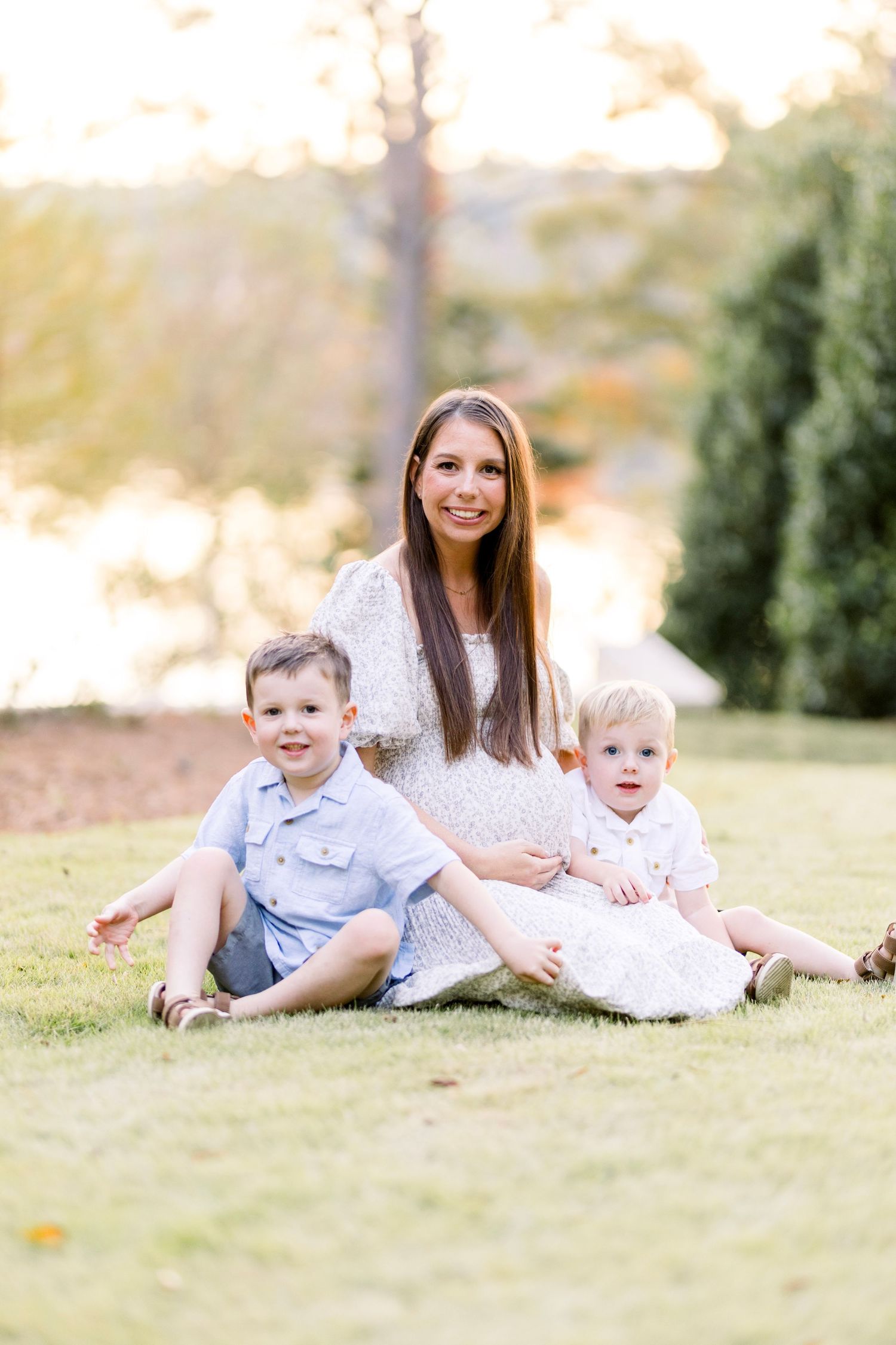A smiling woman sits on green grass with two young children in a sunlit outdoor setting.