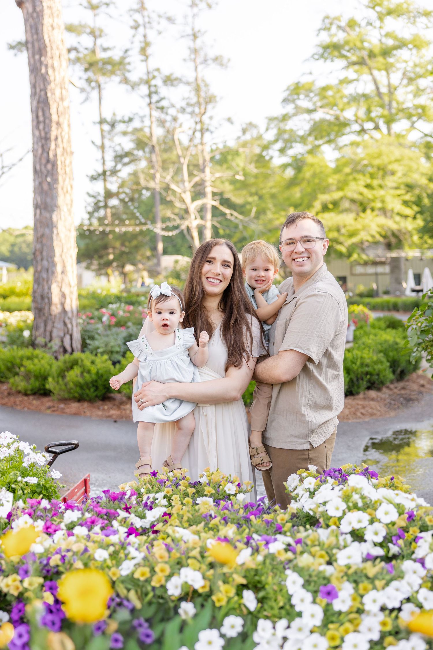 A happy family of four poses among colorful garden flowers, parents holding two young children outdoors.