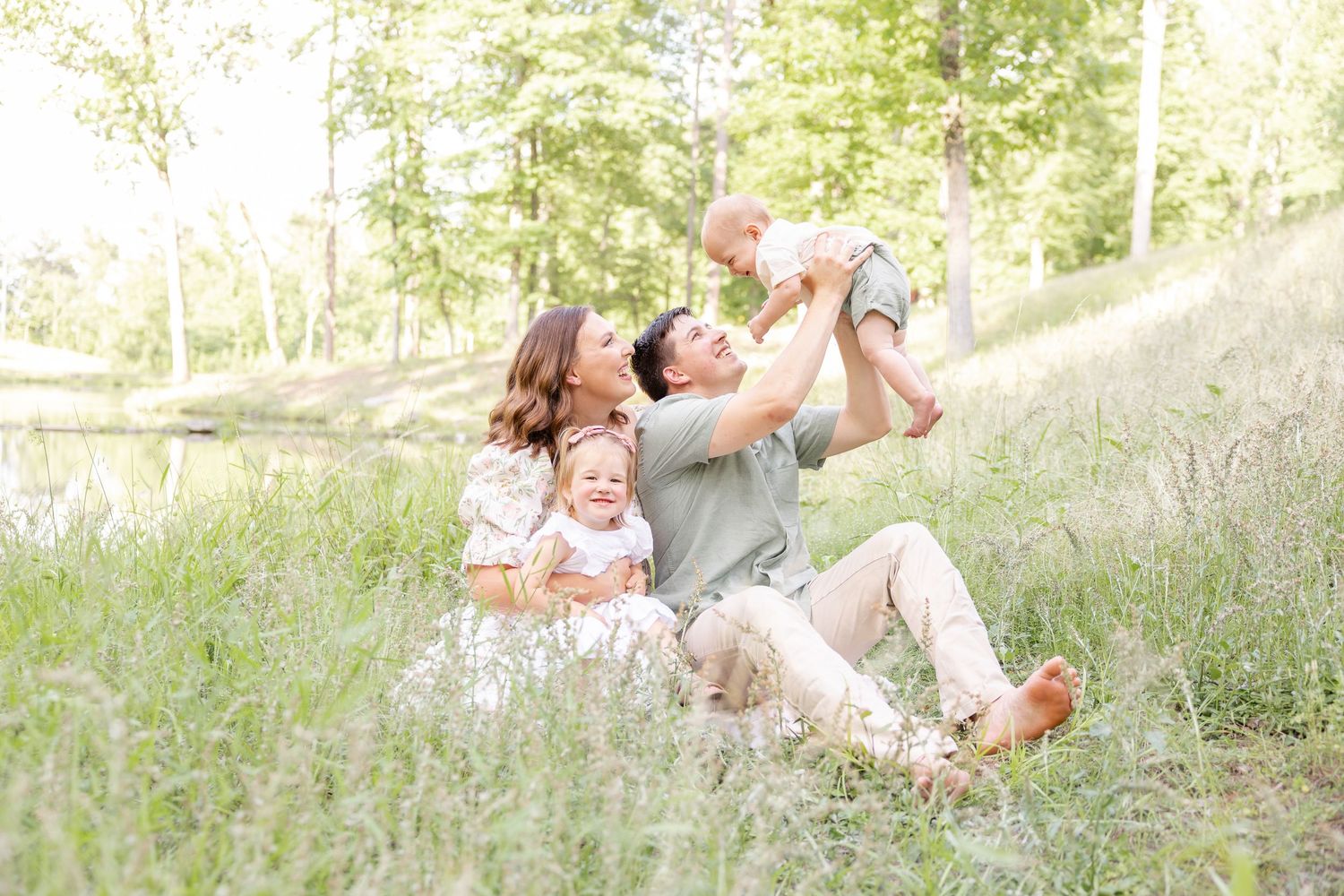 Family relaxing together in sunny meadow with tall grass and trees in background.