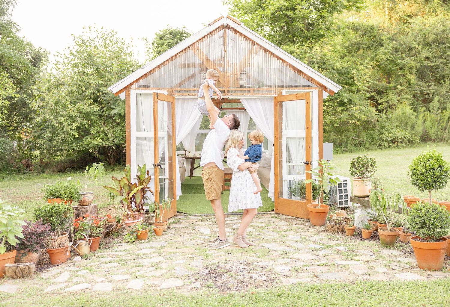 A rustic wooden greenhouse with potted plants lines the stone path entrance while two people stand in the doorway.