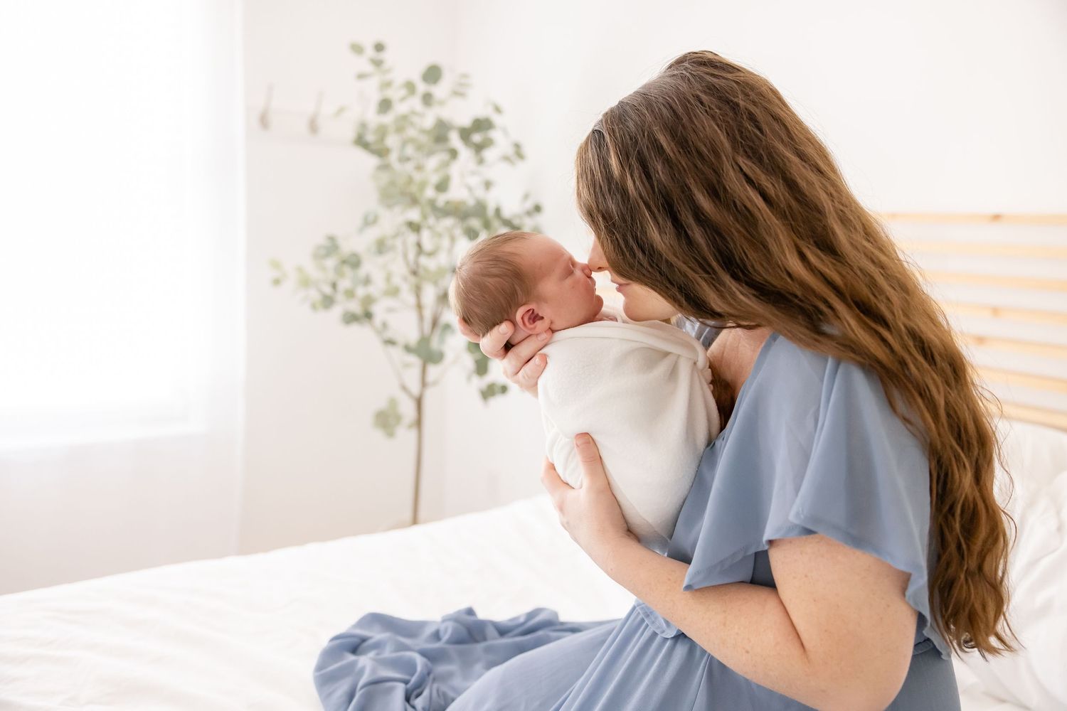 Mother in a beautiful blue dress tenderly holds her sleeping newborn nose to nose while sitting on a bed in bright minimalist bedroom.