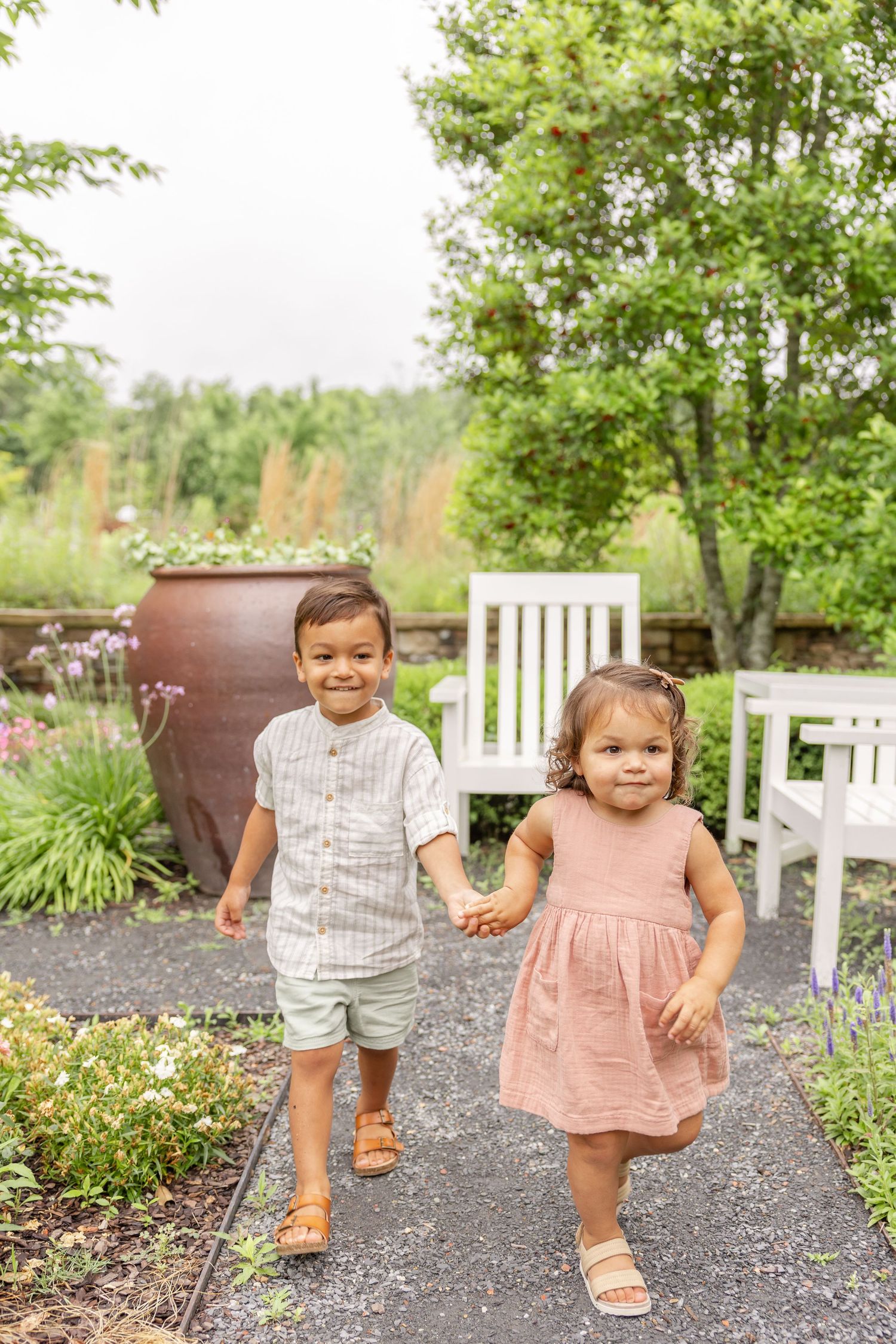 Two toddlers walking, smiling and holding hands on a garden path surrounded by greenery at their family portrait session in Columbus, GA.