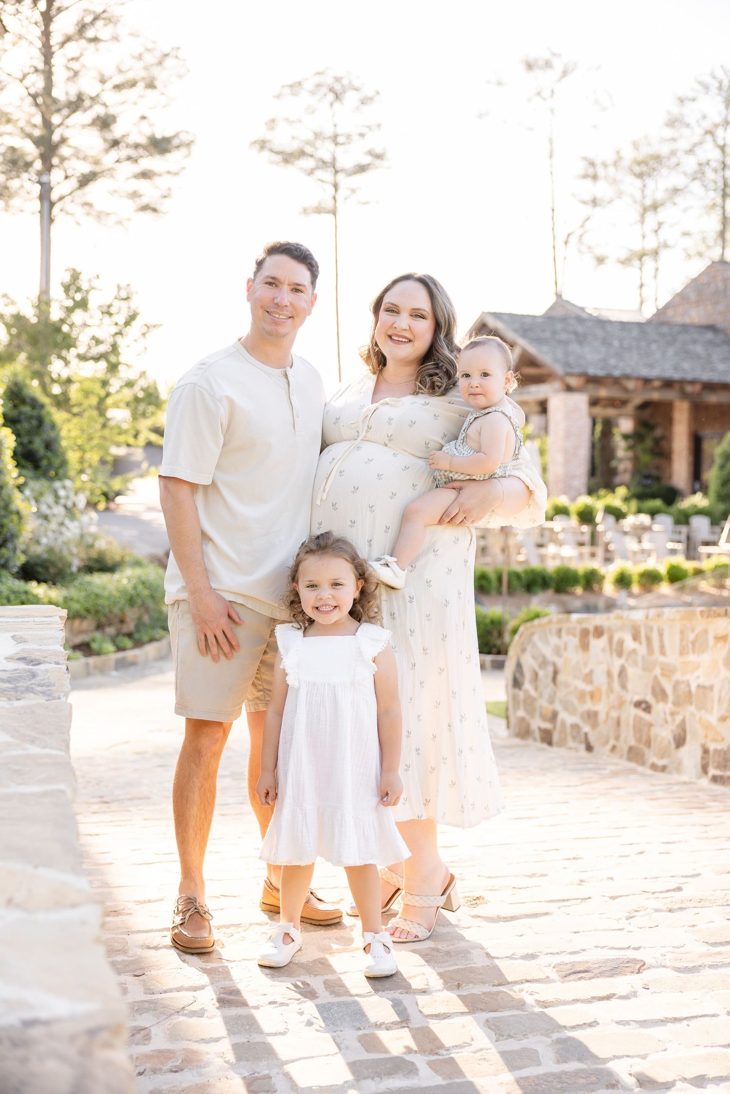 Smiling family of four poses outdoors, pregnant mother holding baby, young daughter in white dress, stone building background.