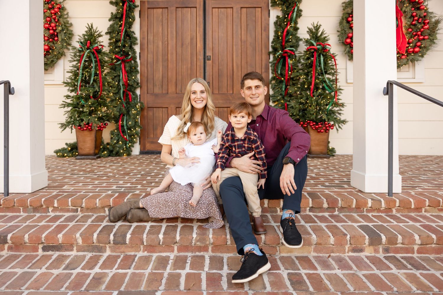 Family poses together on brick stairs decorated with festive holiday garlands and red bows.