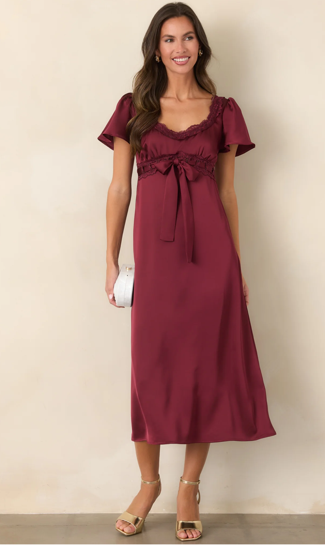 Woman wearing a burgundy satin midi dress with flutter sleeves, lace trim, and bow detail, paired with gold heeled sandals.