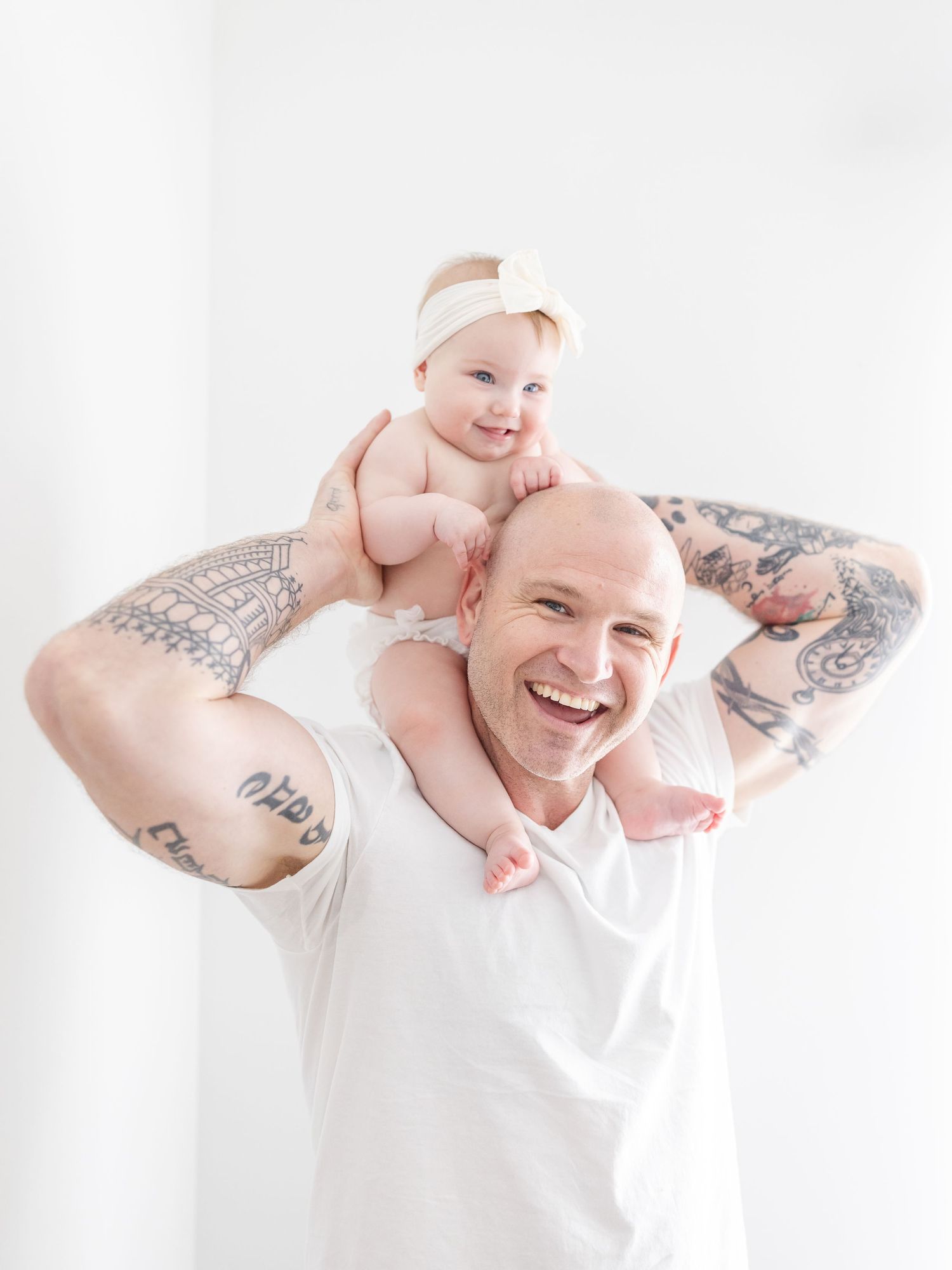 Smiling bald tattooed father holding baby on shoulders against white background.
