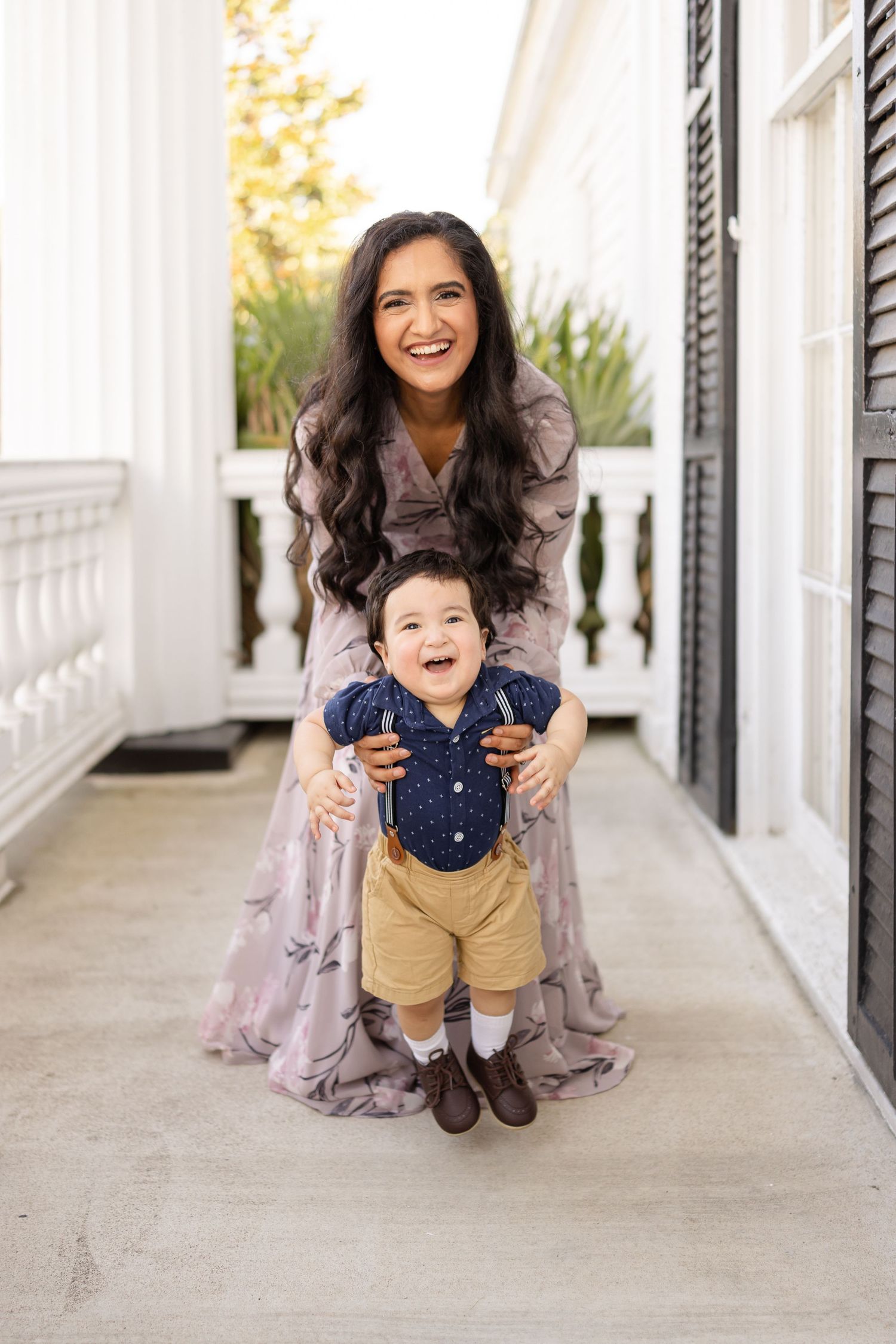 A smiling woman in a floral dress helps a laughing toddler boy stand on a white porch walkway.