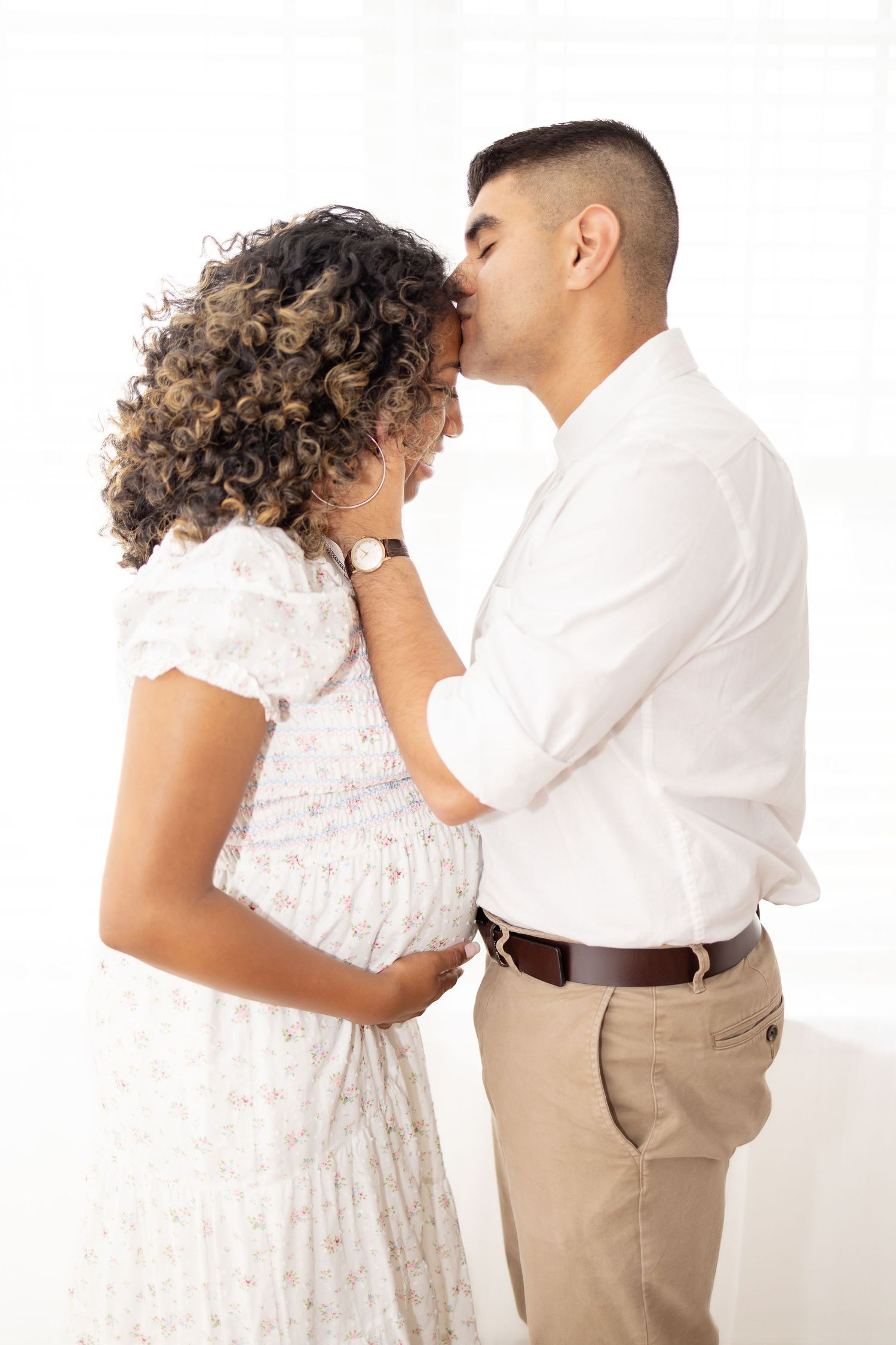 A romantic couple embraces in studio portrait, with one wearing a white floral dress and the other in khaki pants and white shirt.