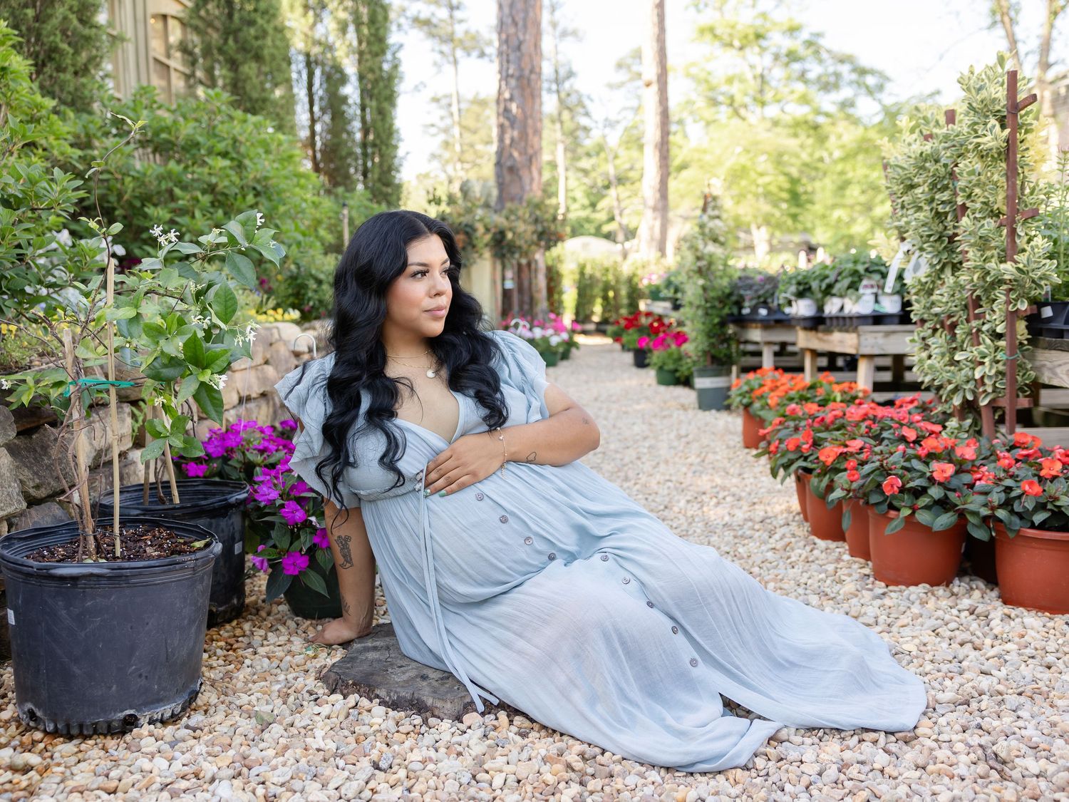 Pregnant woman in blue dress poses gracefully among colorful flowers and potted plants in a lush garden setting.
