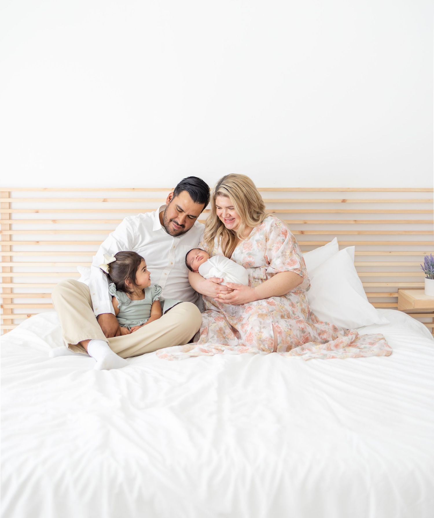 A family sits together on a white bed with wooden headboard, sharing an intimate moment with their newborn baby by Columbus, GA photographer