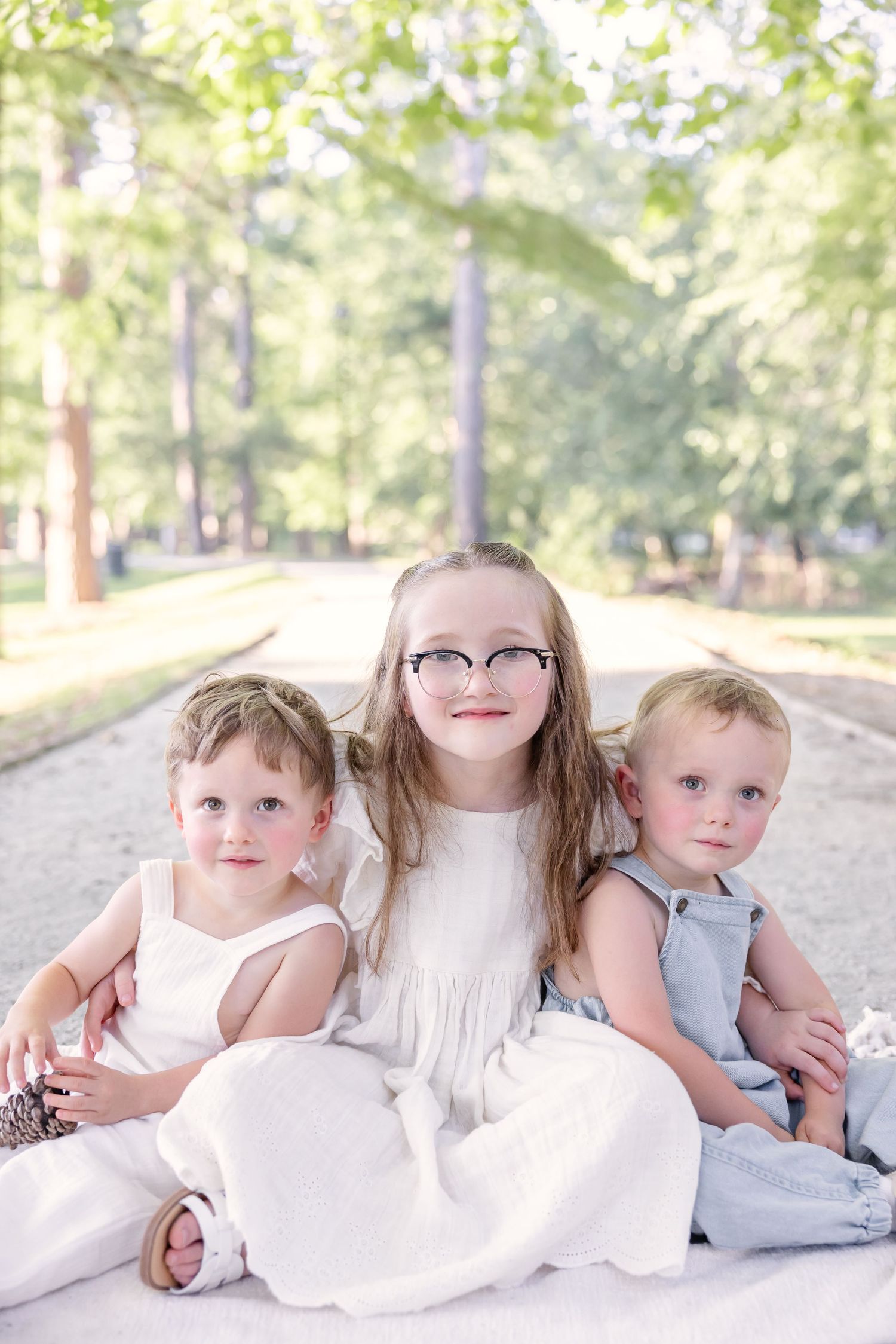 Three young children sitting together outdoors in a park, older girl with glasses flanked by two toddlers in white and blue.