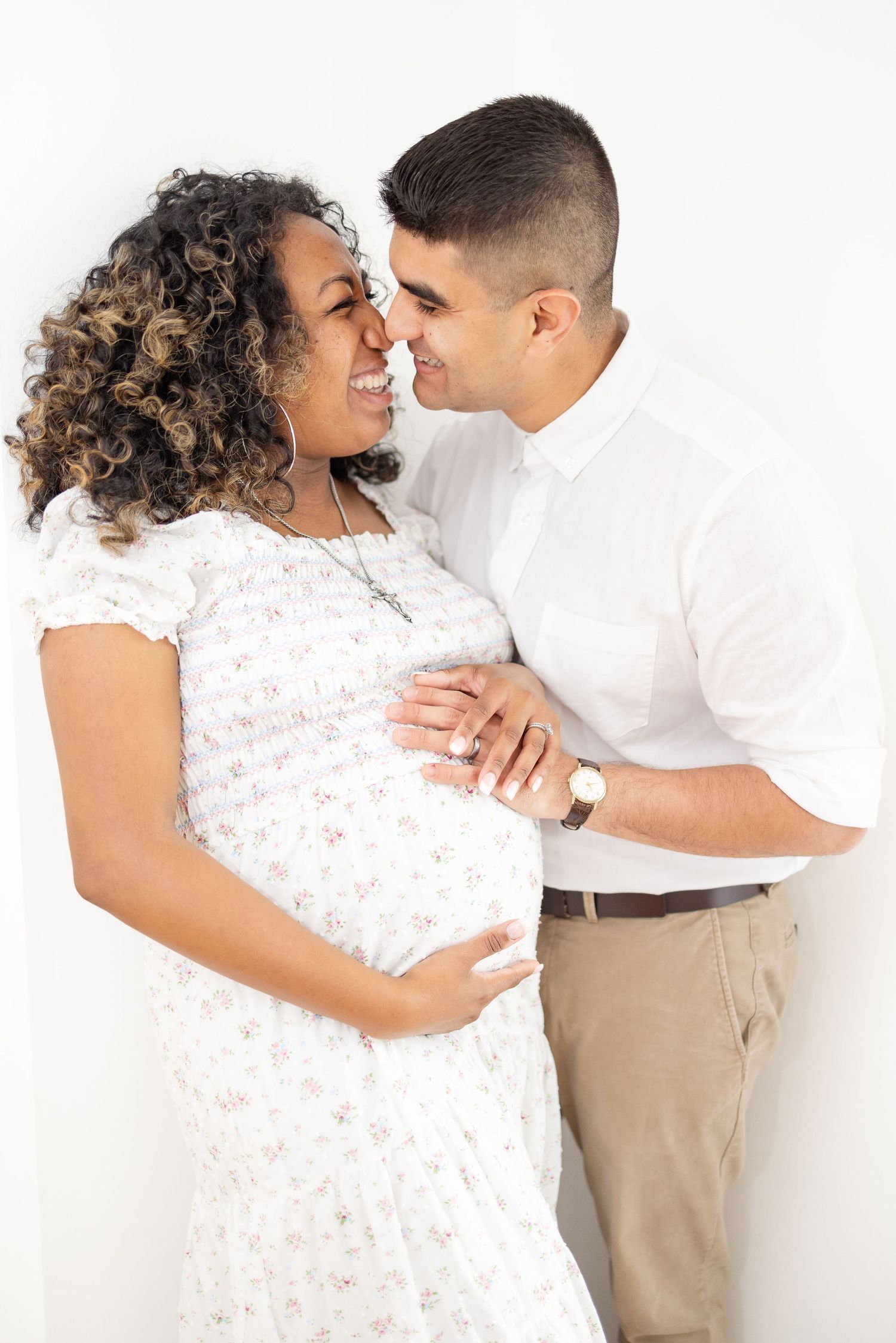 Happy pregnant couple embracing, woman in floral dress cradling baby bump, man in white shirt, white background.
