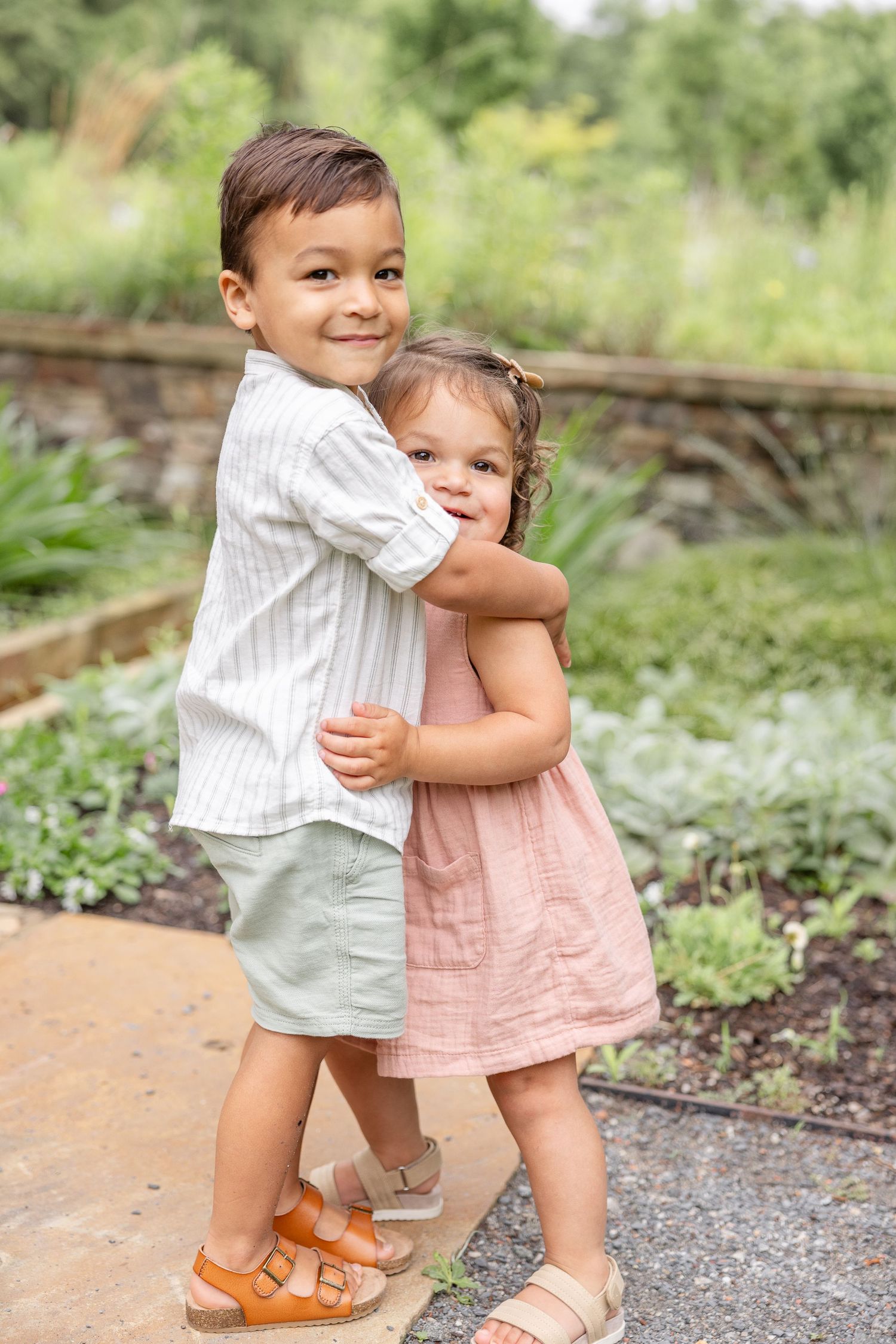 Two young children hugging and smiling on a garden path wearing casual summer outfits during their Columbus, GA family portrait session.