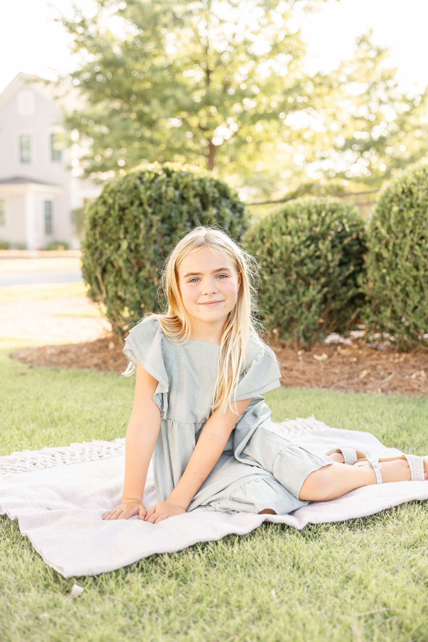 Young blonde girl in sage green dress sitting on blanket in lush green grass during summer family portraits in Columbus, GA.