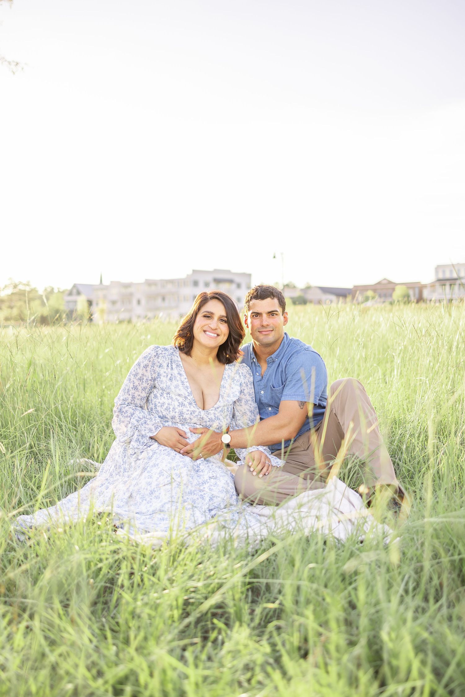 A couple sits together on a blanket in a sunlit meadow of tall grass during their maternity session at golden hour.