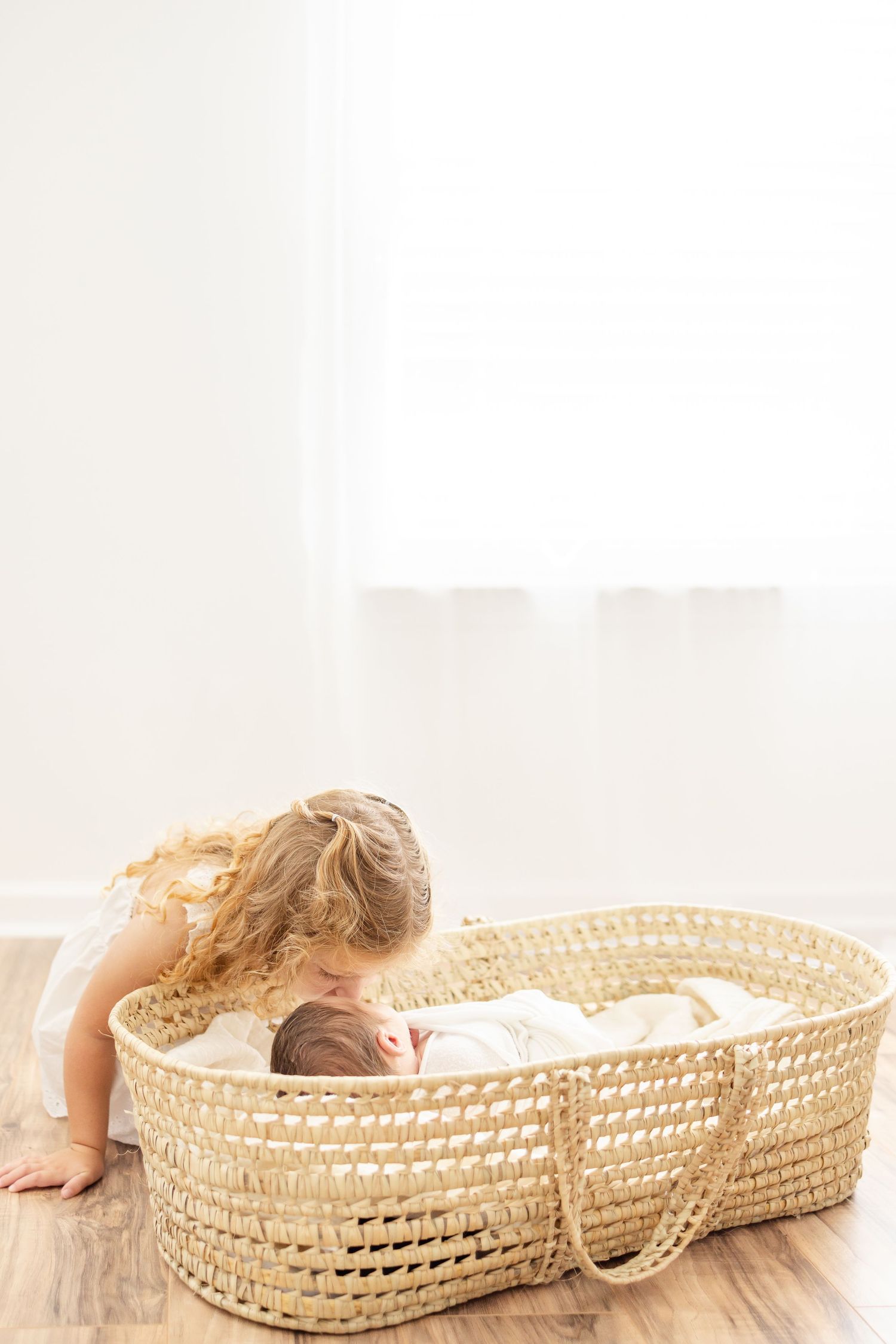A young child with curly hair leans over a woven Moses basket to kiss a sleeping newborn baby.