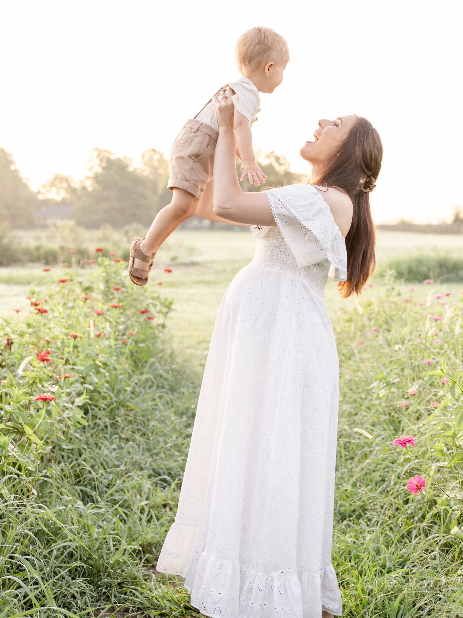 A woman in a flowing white dress lifts a baby in a sunlit field with wildflowers during golden hour.
