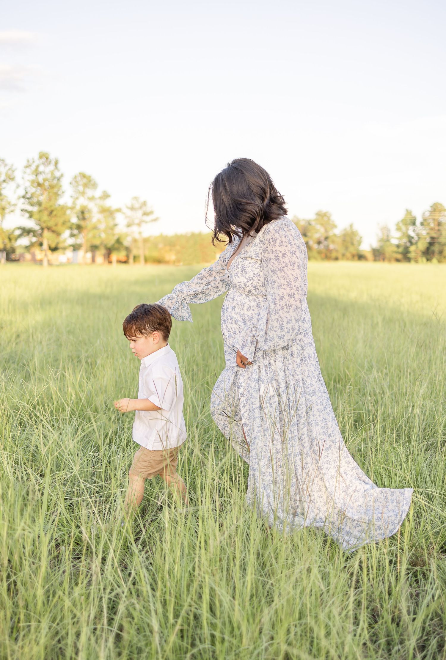 A pregnant mother in a flowing floral dress and her son walk through tall grass in a sunlit meadow during golden hour