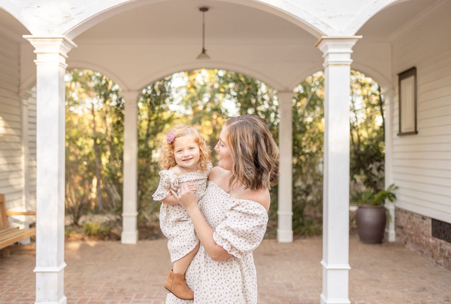 A sunlit porch with white columns frames two people in matching polka dot dresses standing together in a tender moment.