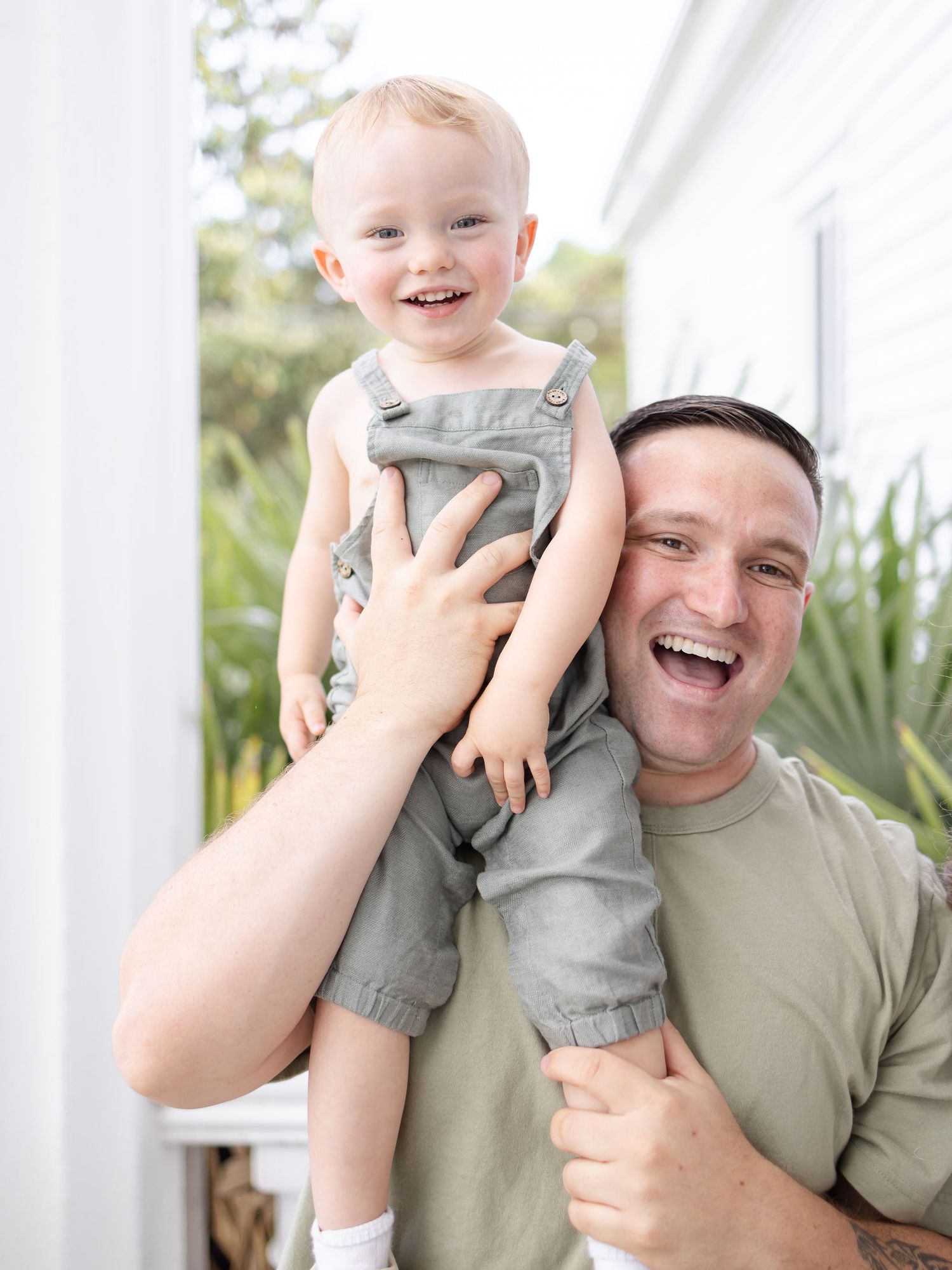 Smiling dad holding his laughing son in green overalls on his shoulders outdoors during spring family portraits in Columbus, GA.