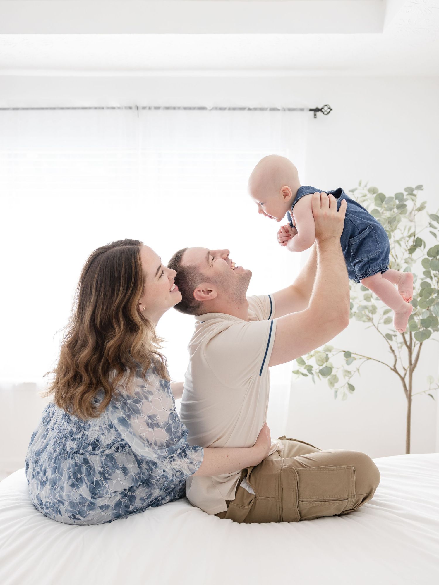 A happy young family sits on a white bed, with dad lifting smiling baby in the air while mom looks on lovingly.