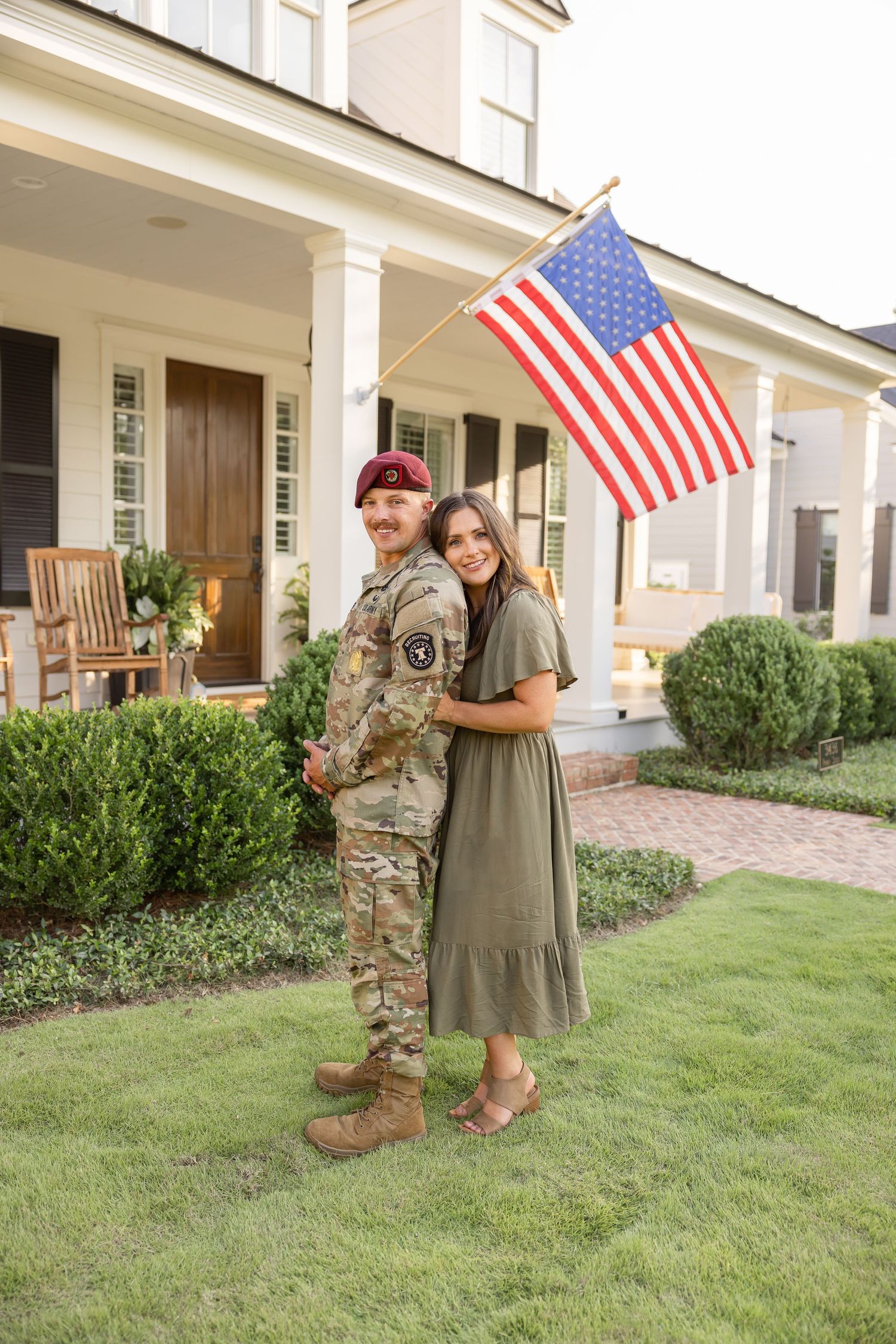 Military service member and his wife embrace on front lawn of home with American flag displayed on porch by Columbus, GA family photographer