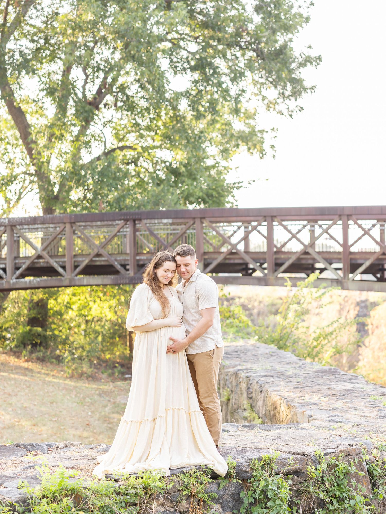 A couple poses tenderly on stone steps near a wooden bridge, surrounded by lush greenery in warm golden light.