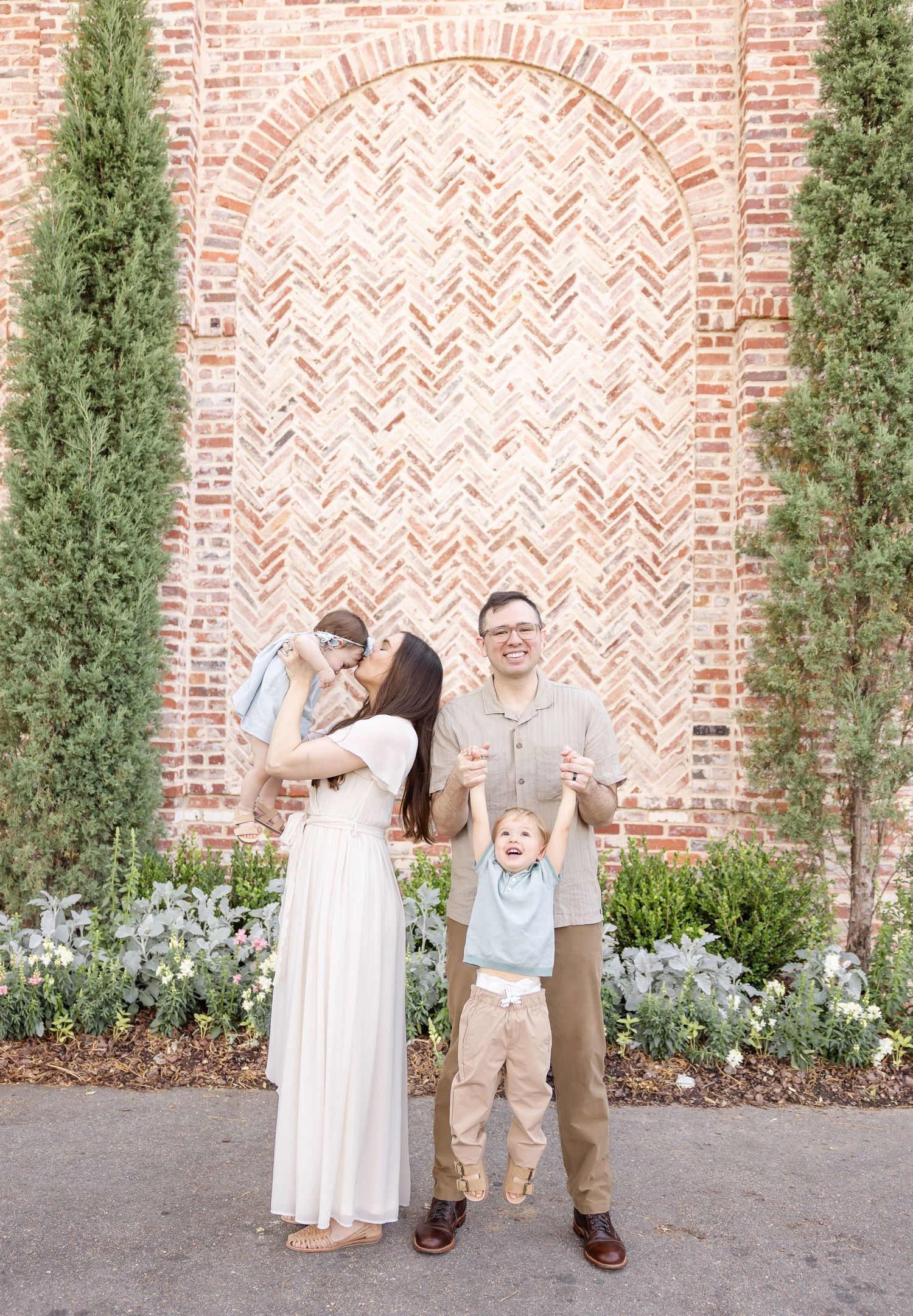Family of four posing in front of a herringbone brick wall with cypress trees and flowers.