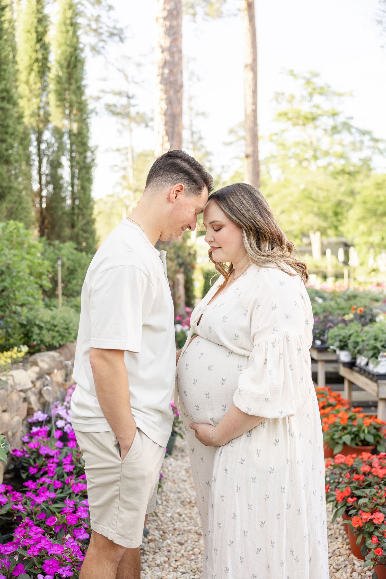 A couple in white outfits share a tender moment in a flower garden, with the woman cradling her pregnant belly.