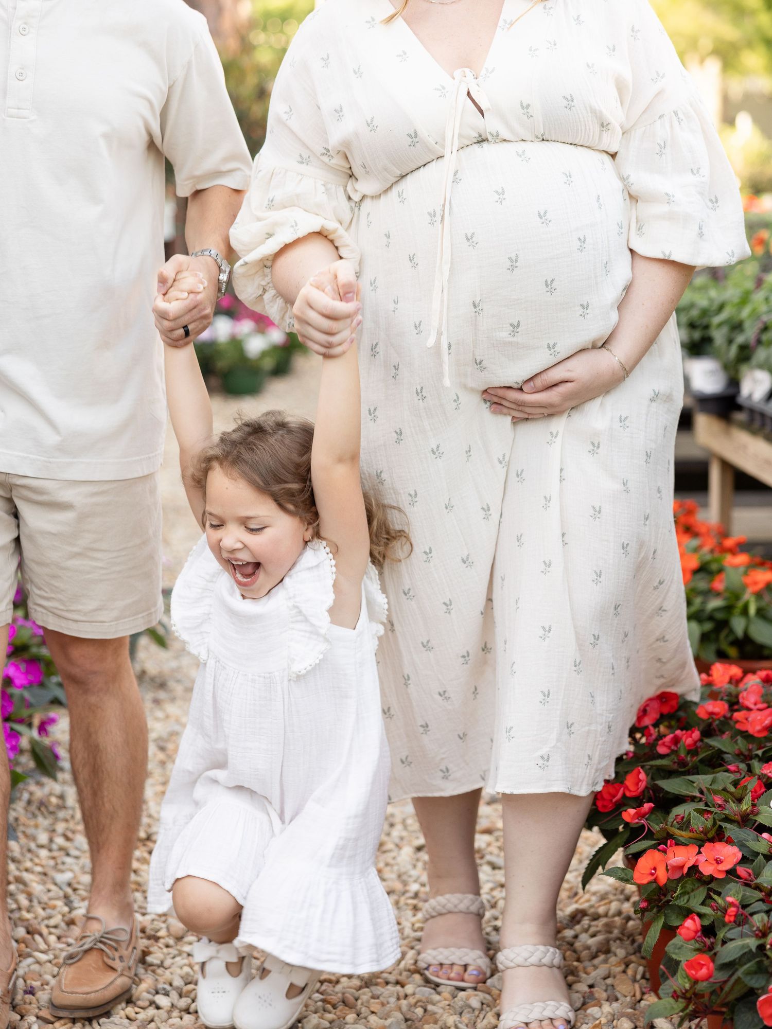 Pregnant woman in floral dress with toddler girl laughing, held by parents, surrounded by colorful flowers for outdoor maternity portraits.