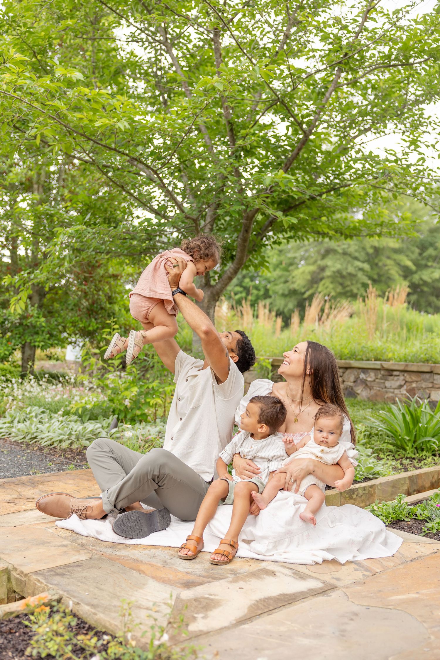 Family relaxes together on white blanket in lush garden setting during outdoor photo session in Columbus, GA.