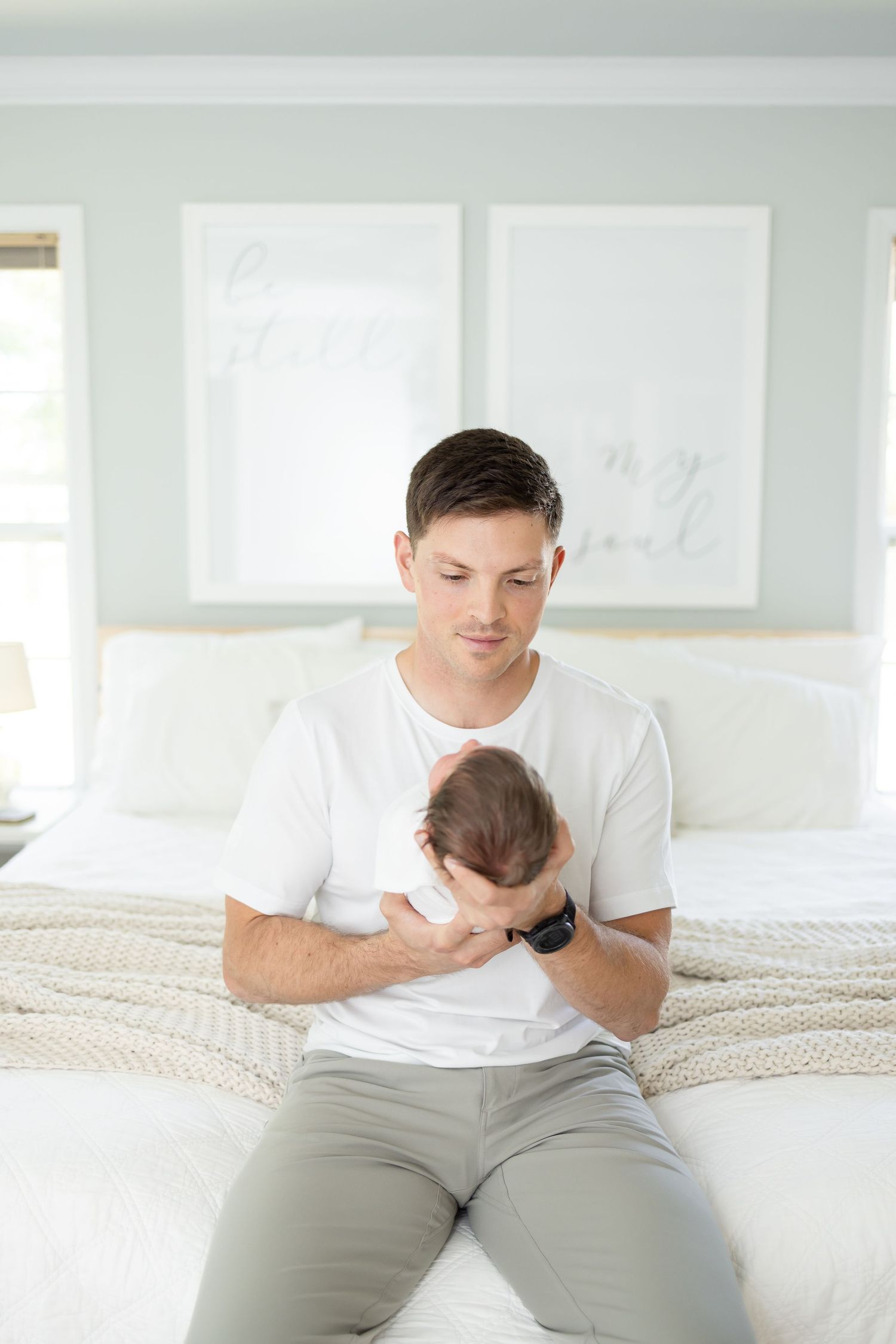 A father in a white t-shirt and gray pants sits on a bed holding his newborn daughter during his in-home lifestyle newborn portrait session.