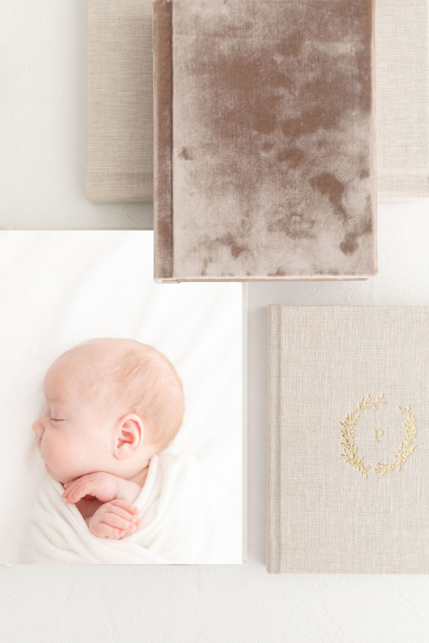 Newborn baby sleeping peacefully in white wrap beside elegant linen and velvet photo albums with gold monogram.