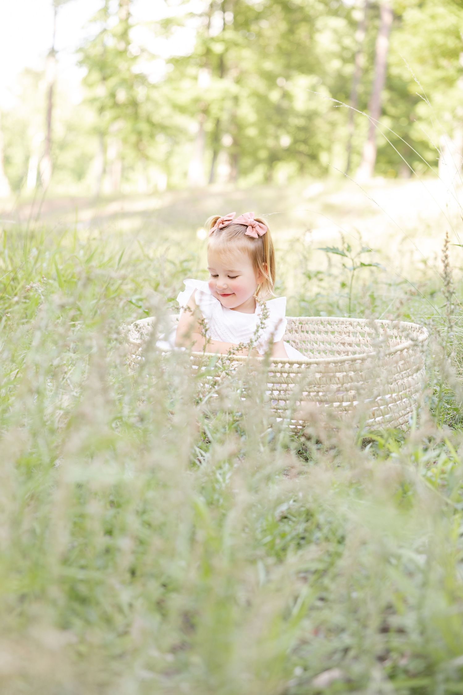 A child in a white dress sits in a woven basket surrounded by tall green grass by Columbus, GA photographer.
