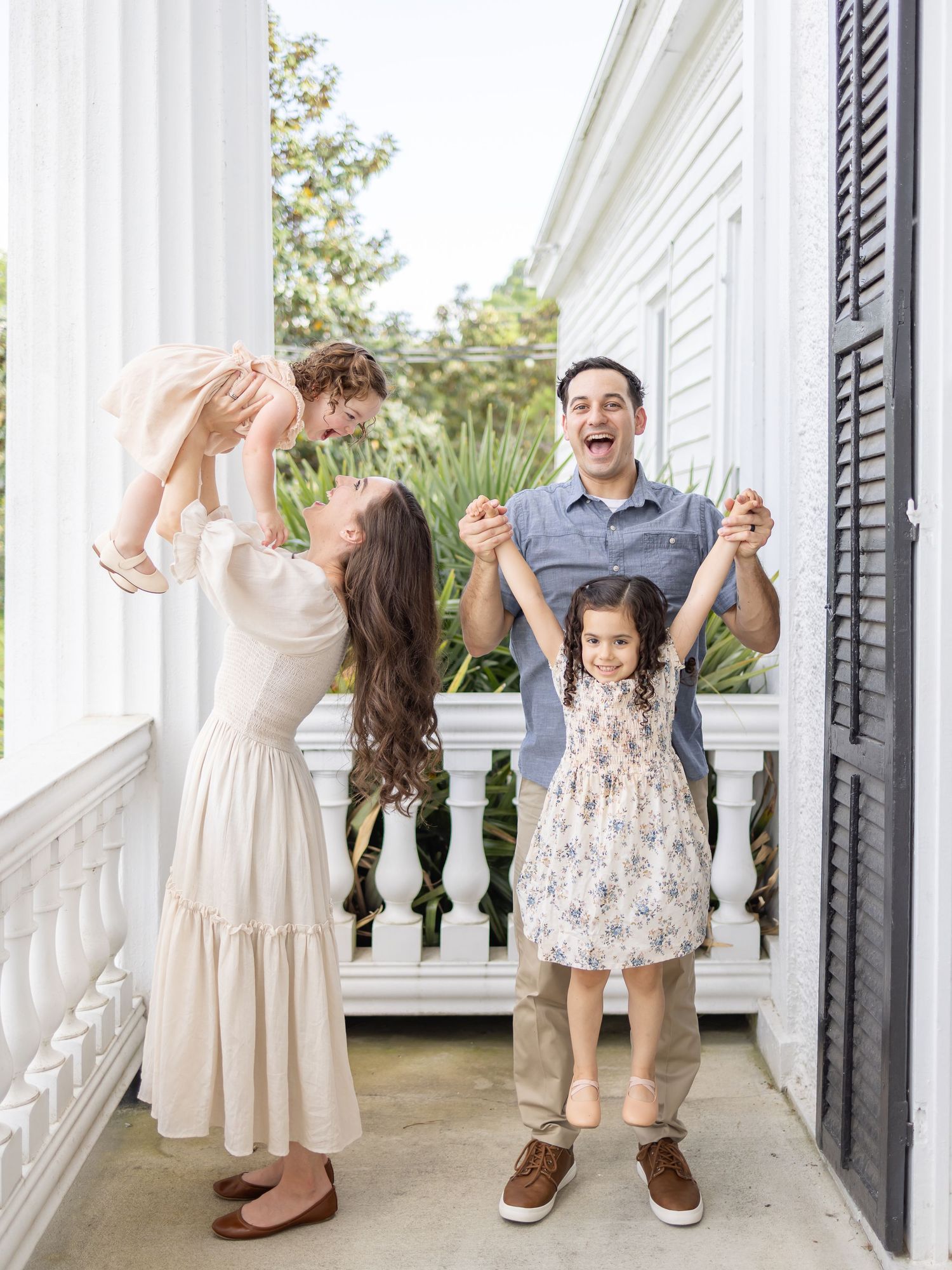 A family enjoys a playful moment together on a white columned porch during a bright summer day.