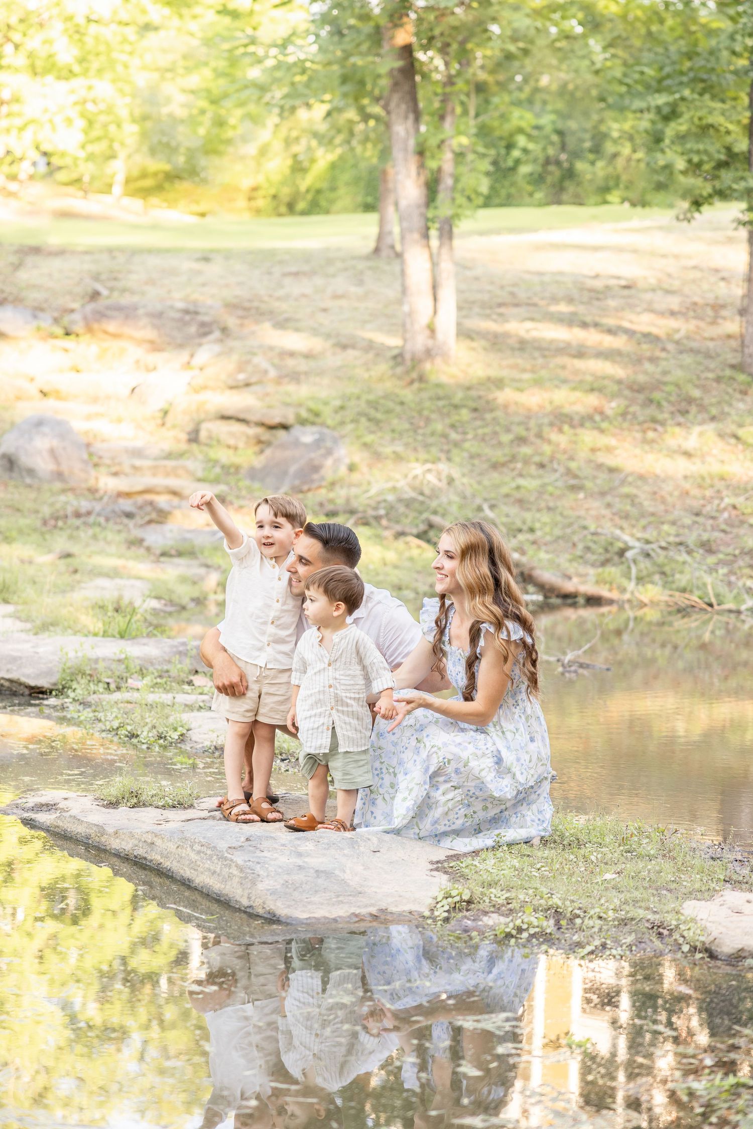 A family spends time together near a tranquil pond in a sunlit wooded area on a summer afternoon.