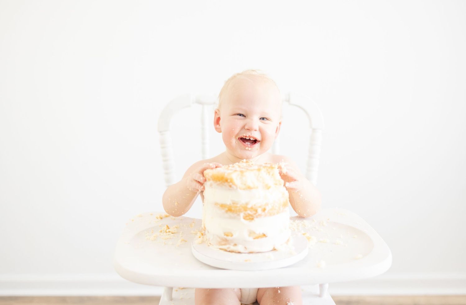 Laughing baby sitting in white high chair with frosted cake during first birthday celebration photoshoot by Columbus, GA photographer.