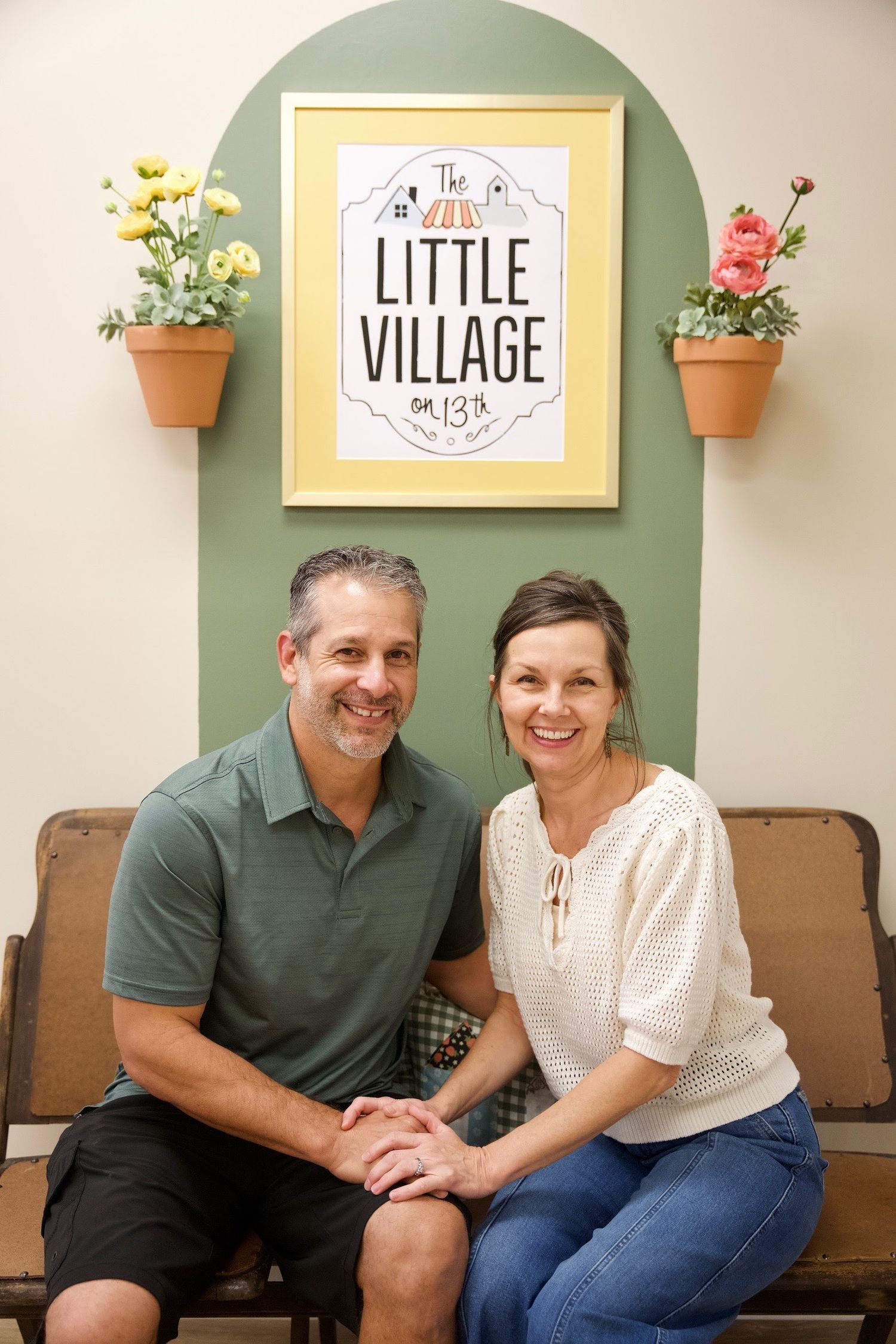 A cozy interior scene with potted flowers and a framed 'Little Village' sign on a sage green wall above a brown bench.