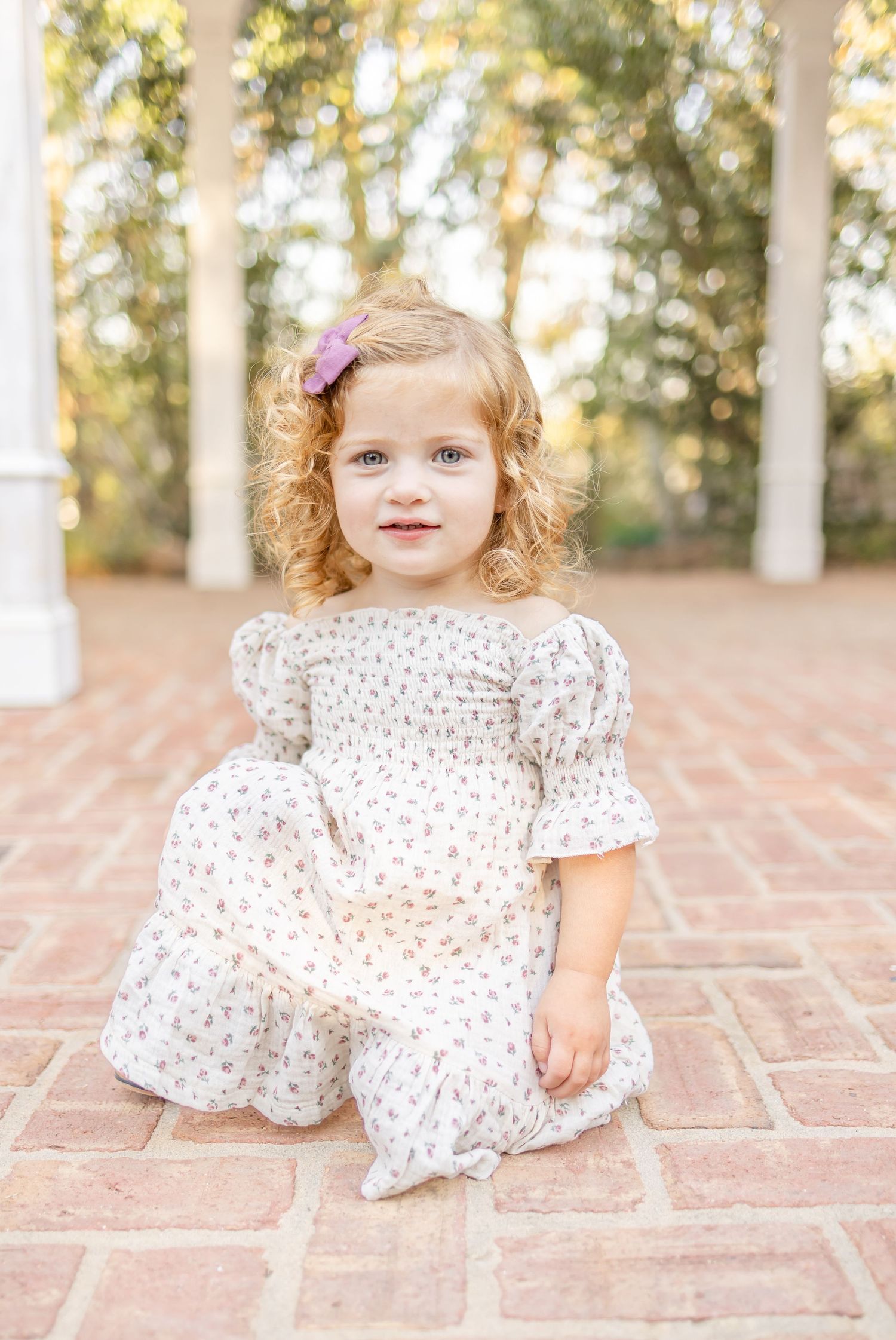 Child in floral print dress with puff sleeves and pink hair bow sits on brick patio in sunlit outdoor setting.
