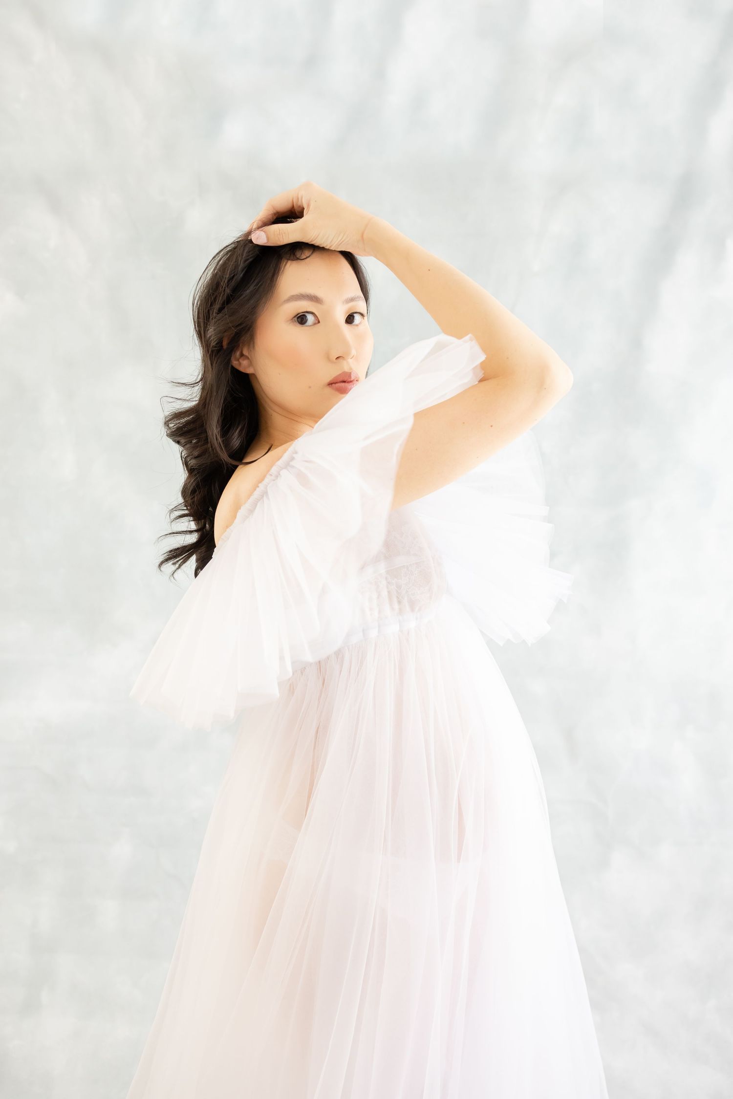 A pregnant mother in a flowing white tulle dress poses gracefully against a light background at her studio maternity session in Columbus GA