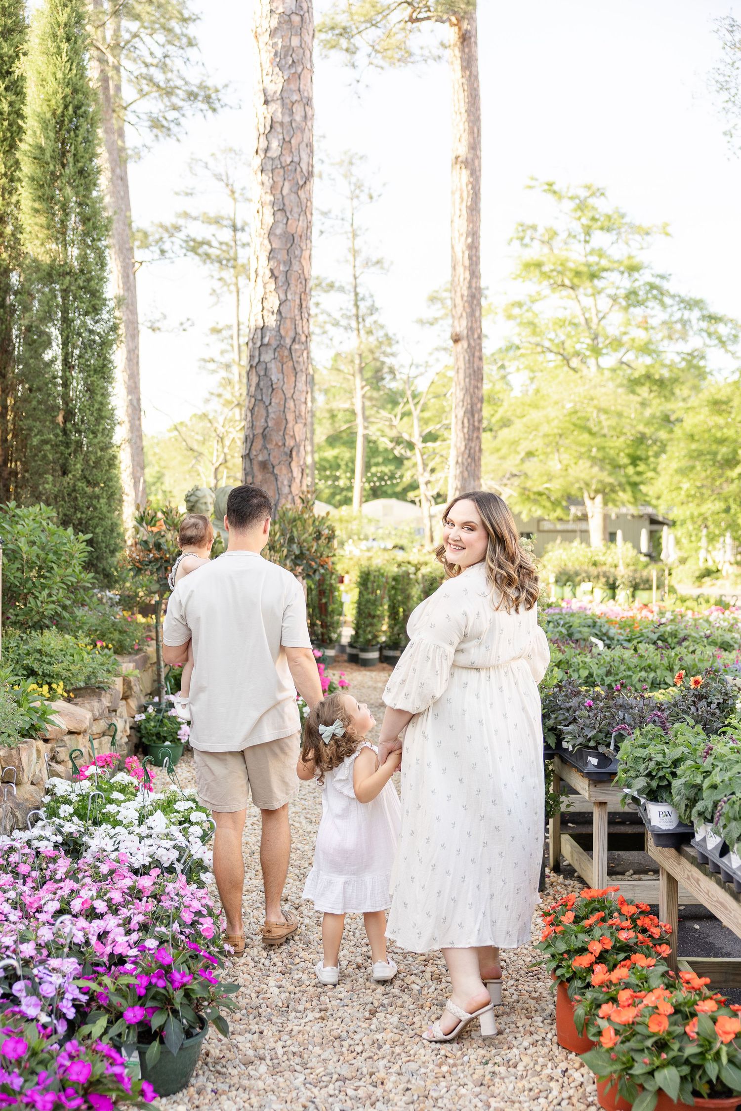 Pregnant woman in white dress smiles back at camera while family walks through a lush garden nursery.