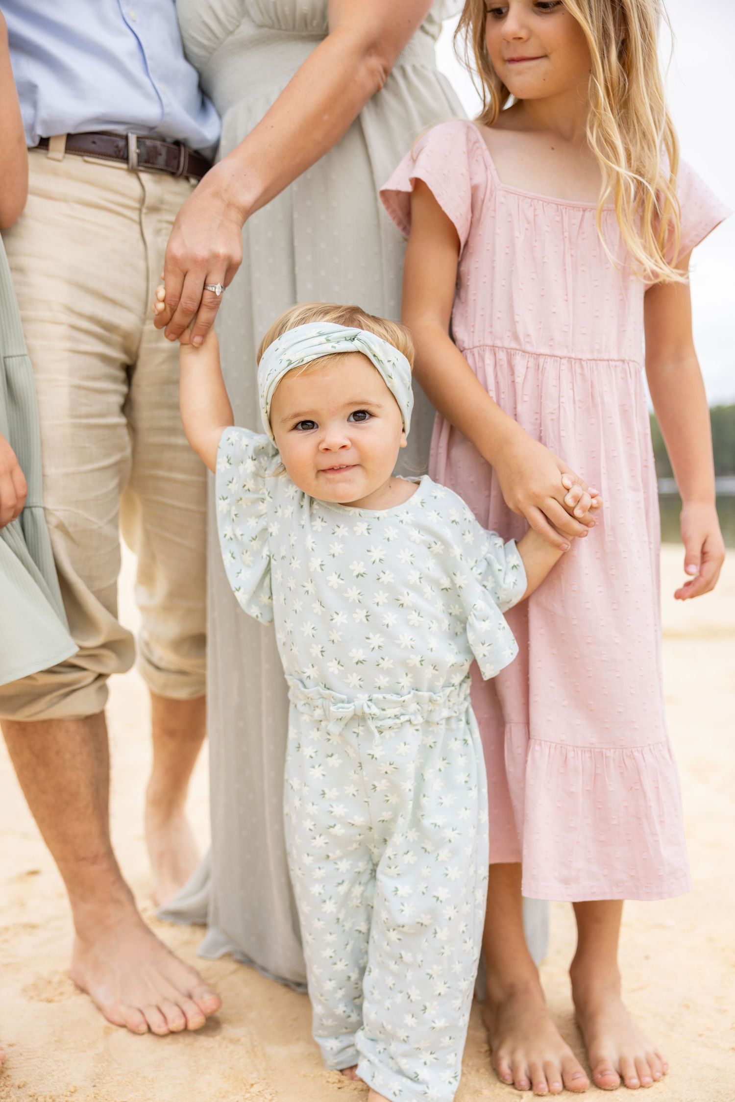 Family wearing coordinated pastel outfits stands barefoot together on light colored surface.