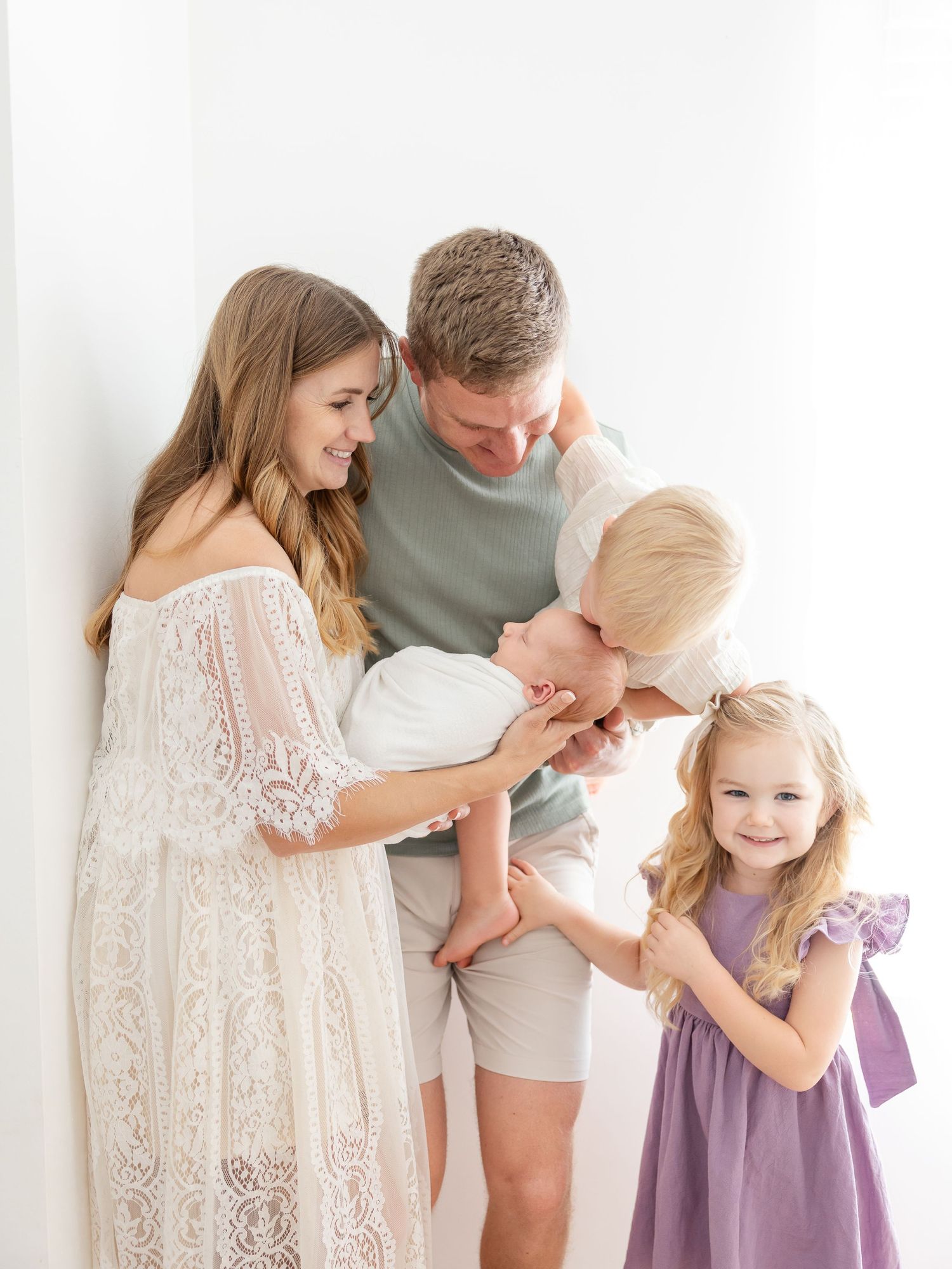 A loving family of four with a newborn poses against a white wall in soft, neutral tones for a studio portrait.